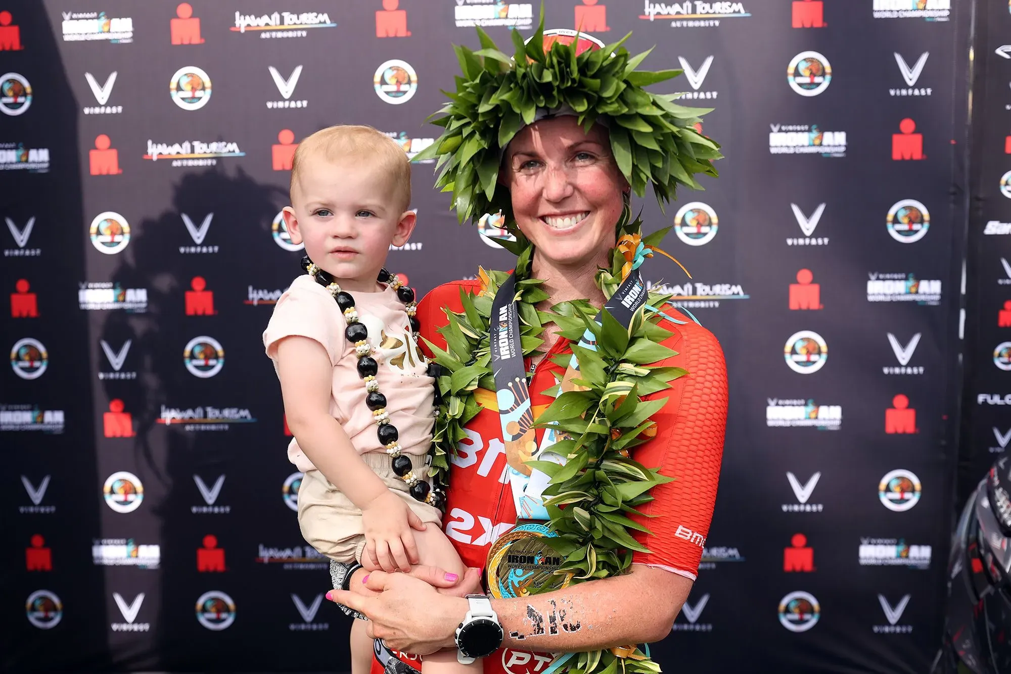 امرأة تحمل طفلاً وتبتسم بفخر بعد الفوز بسباق، ترتدي ميدالية وزهورًا، تعكس قوة الرياضيين الأمهات في مواجهة التحديات.