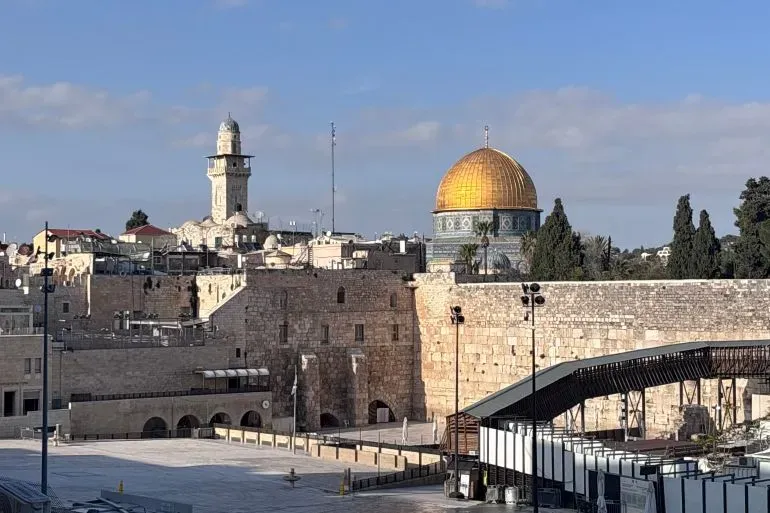 مشهد لمدينة القدس القديمة يظهر قبة الصخرة والمآذن، مع شوارع فارغة ومحلات مغلقة، مما يعكس تأثير الأوضاع السياسية على الحياة اليومية.