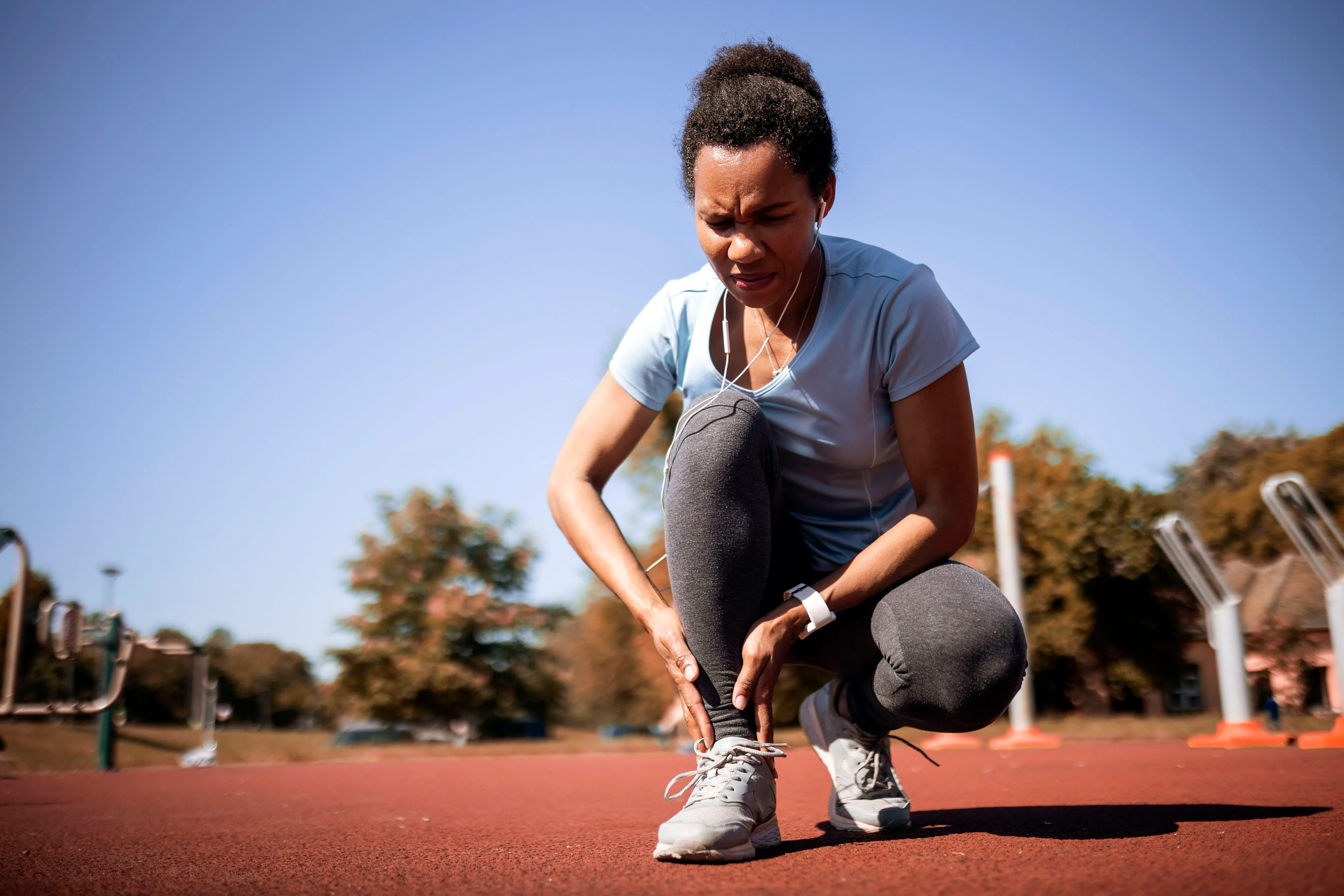 امرأة ترتدي ملابس رياضية، تجلس على الأرض وتلمس كاحلها، مع تعبير عن الألم، في بيئة رياضية. تشير الصورة إلى أهمية حركة الكاحل لصحة الجسم.