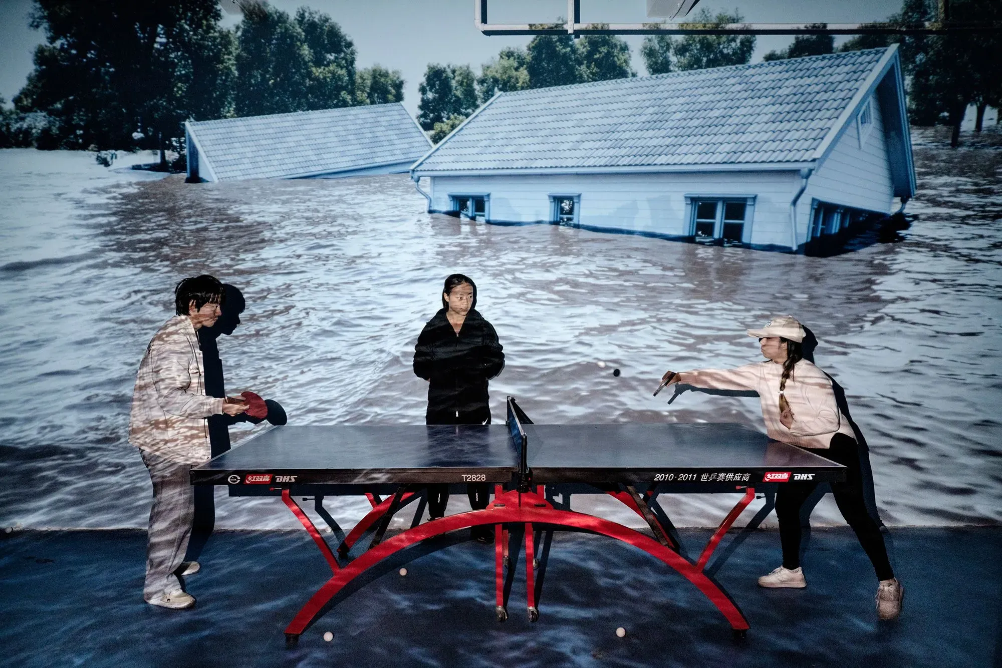 صورة تظهر لاعبي تنس الطاولة في مكان مغمور بالمياه، مع منازل غارقة في الخلفية، تعكس تأثيرات تغير المناخ على المجتمعات.