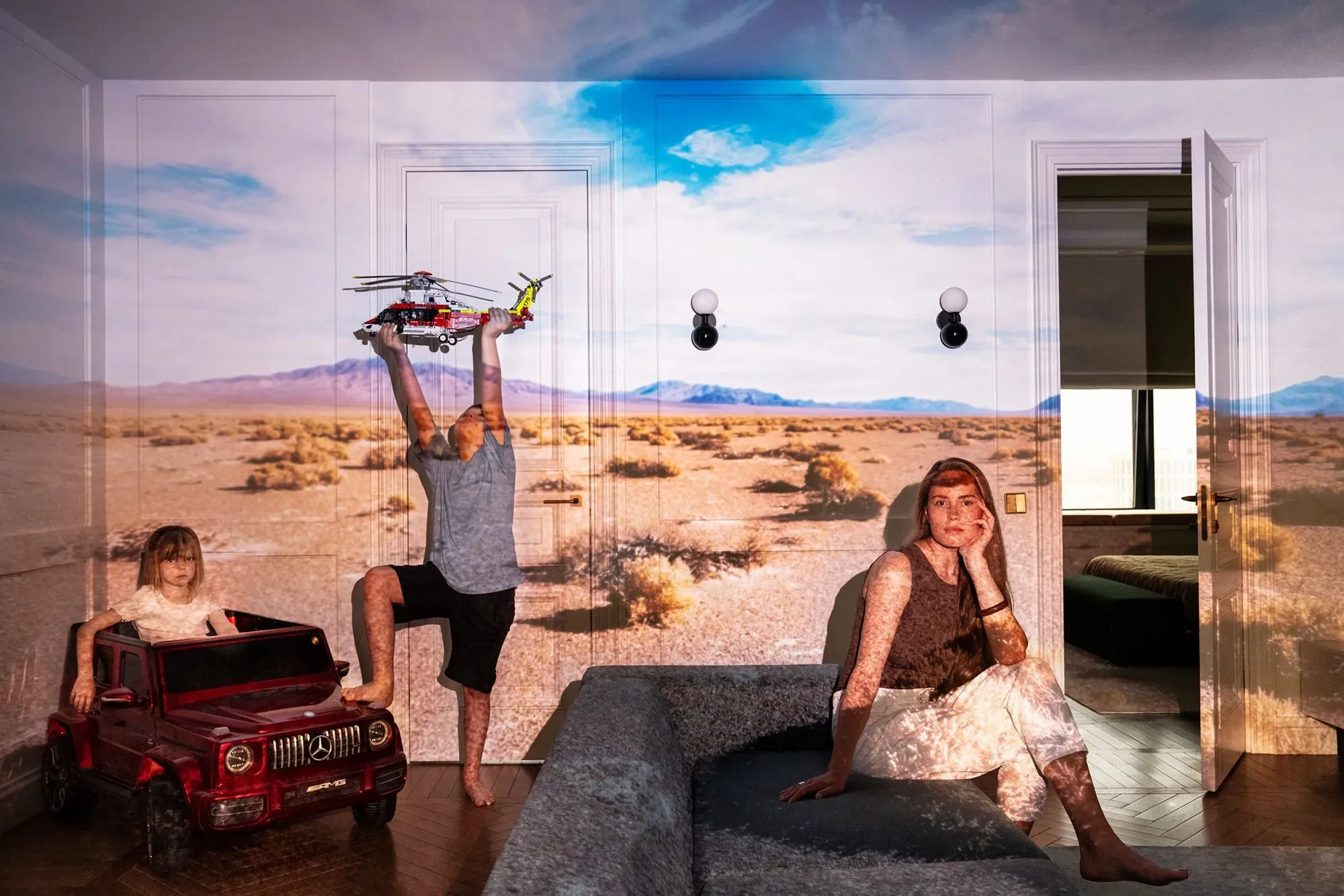 صورة عائلية تظهر طفلين يلعبان في غرفة معيشة، مع خلفية مناظر طبيعية صحراوية مدمجة، تعكس تأثير الاحتباس الحراري.