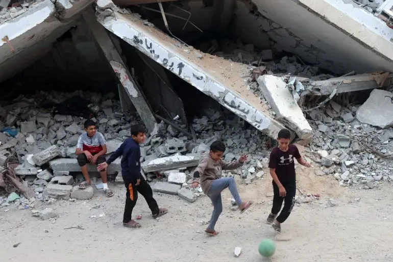 أطفال يلعبون كرة القدم أمام أنقاض مبنى مدمر في غزة، مما يعكس آثار الصراع وأزمة السكن في المنطقة.