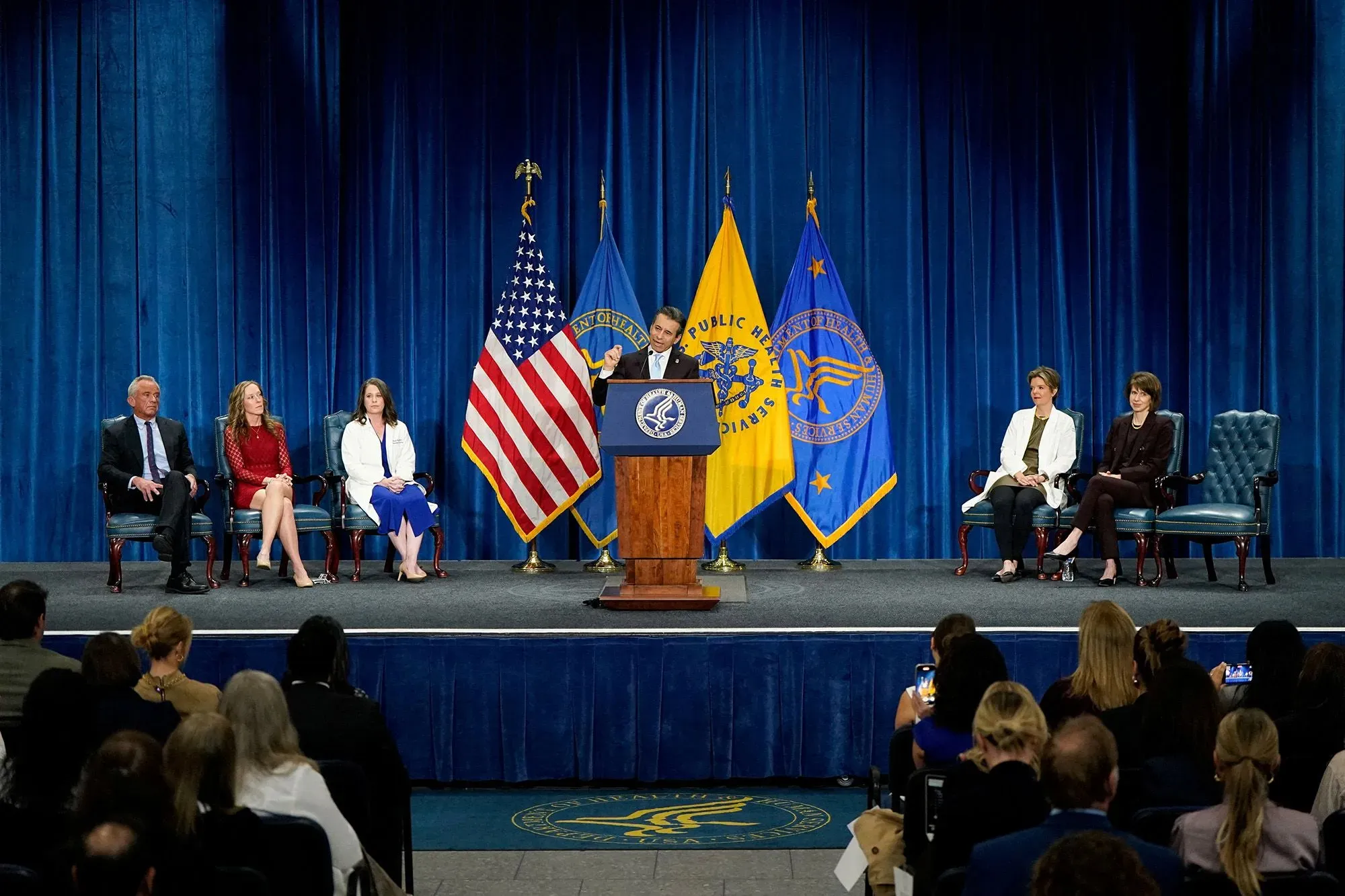 وزير الصحة الأمريكي يتحدث في مؤتمر صحفي عن إزالة التحذير من العلاج الهرموني، مع وجود جمهور وأعلام خلفه.