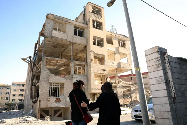 مبنى مدمر جزئياً في إيران، يظهر آثار الحرب، مع امرأتين تسيران بجانب المبنى، مما يعكس تأثير النزاع على الحياة اليومية.