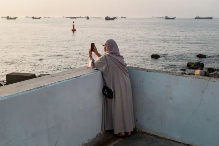 امرأة ترتدي عباءة تقف على حافة رصيف، تلتقط صورة للبحر والسفن في مضيق هرمز، في ظل تصاعد التوترات الجيوسياسية.