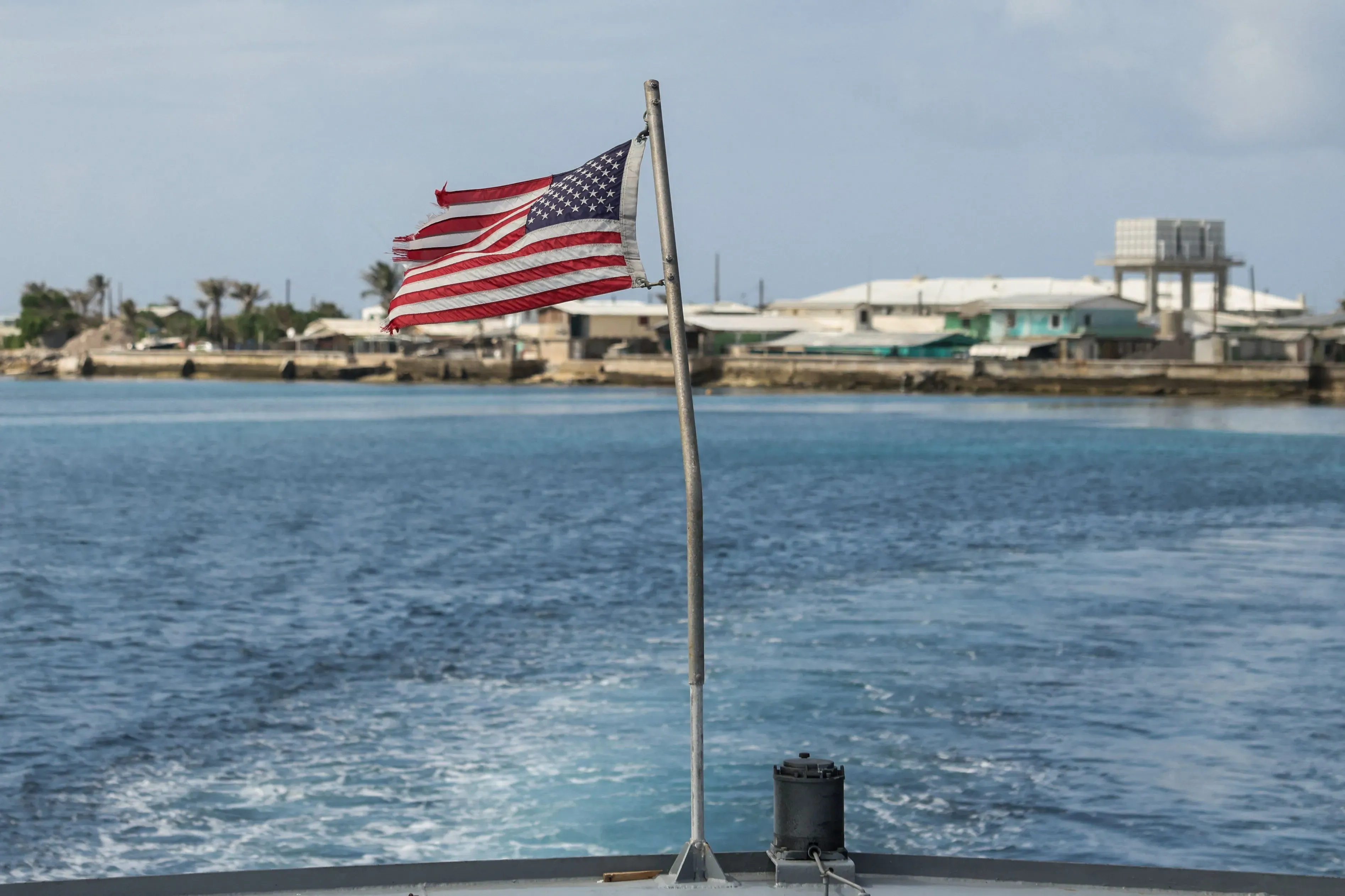 علم الولايات المتحدة ممزق يرفرف فوق مياه جزر مارشال، مع خلفية تظهر المباني الساحلية، مما يعكس آثار التجارب النووية.