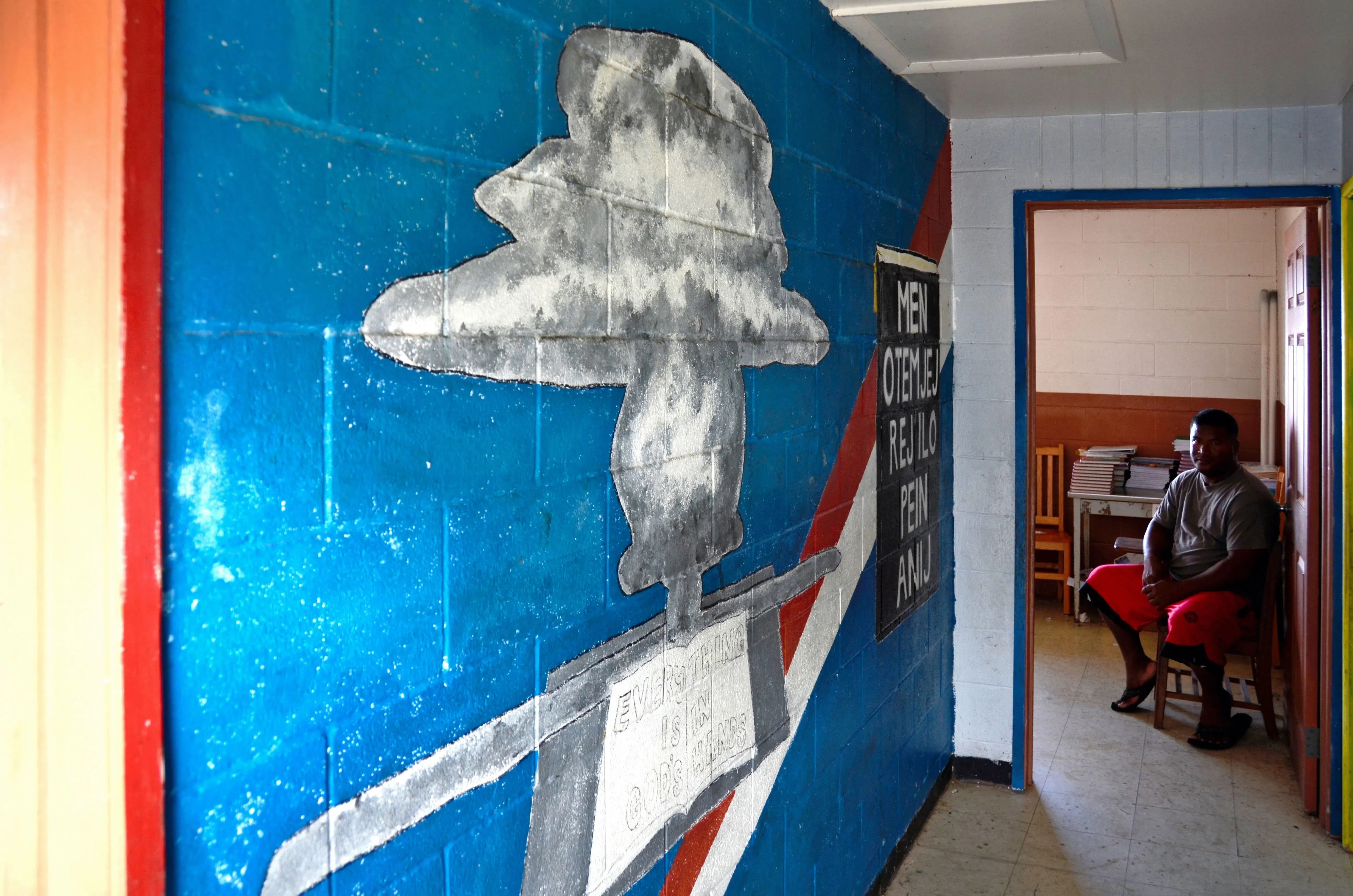 رسم جداري يظهر سحابة انفجار نووي على جدار ملون، مع رجل يجلس بجوار المدخل في جزر مارشال، يعكس آثار التجارب النووية.