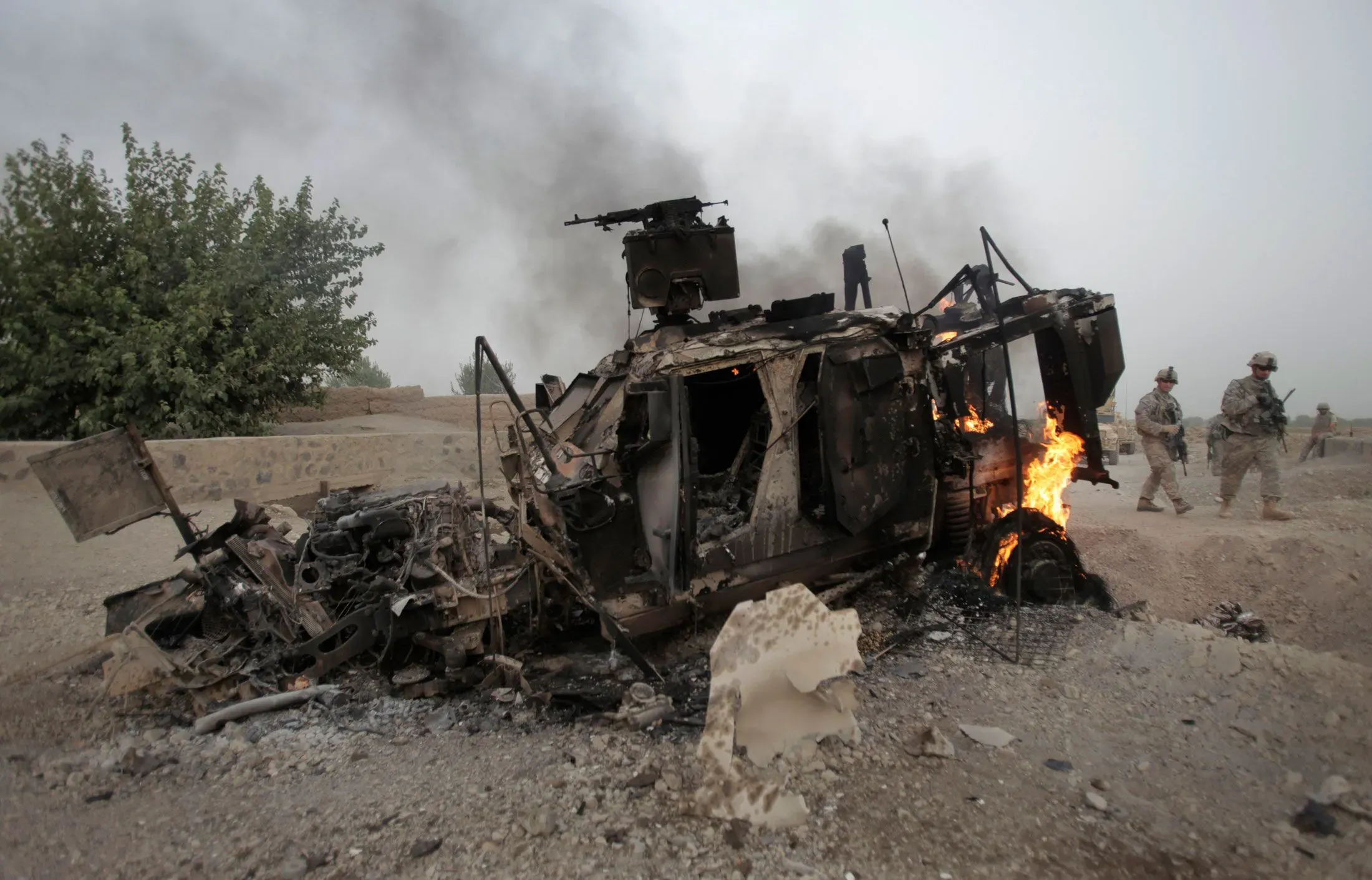 حطام مركبة عسكرية محترقة في منطقة قتال، مع جنود أمريكيين يتجولون في الخلفية، مما يعكس التهديد المتزايد للطائرات بدون طيار.