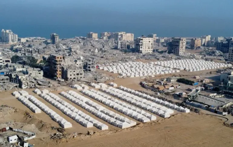 تظهر الصورة مشهدًا لمدينة غزة المدمرة، مع خيام بيضاء مرتبة في صفوف، تعكس الأزمة الإنسانية بعد الهجمات الإسرائيلية.