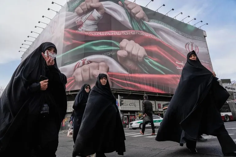 نساء يرتدين الحجاب الأسود يسيرون في الشارع أمام لافتة ضخمة تُظهر أيادي مرفوعة تحمل الأعلام الإيرانية، تعبيرًا عن الهوية الوطنية.