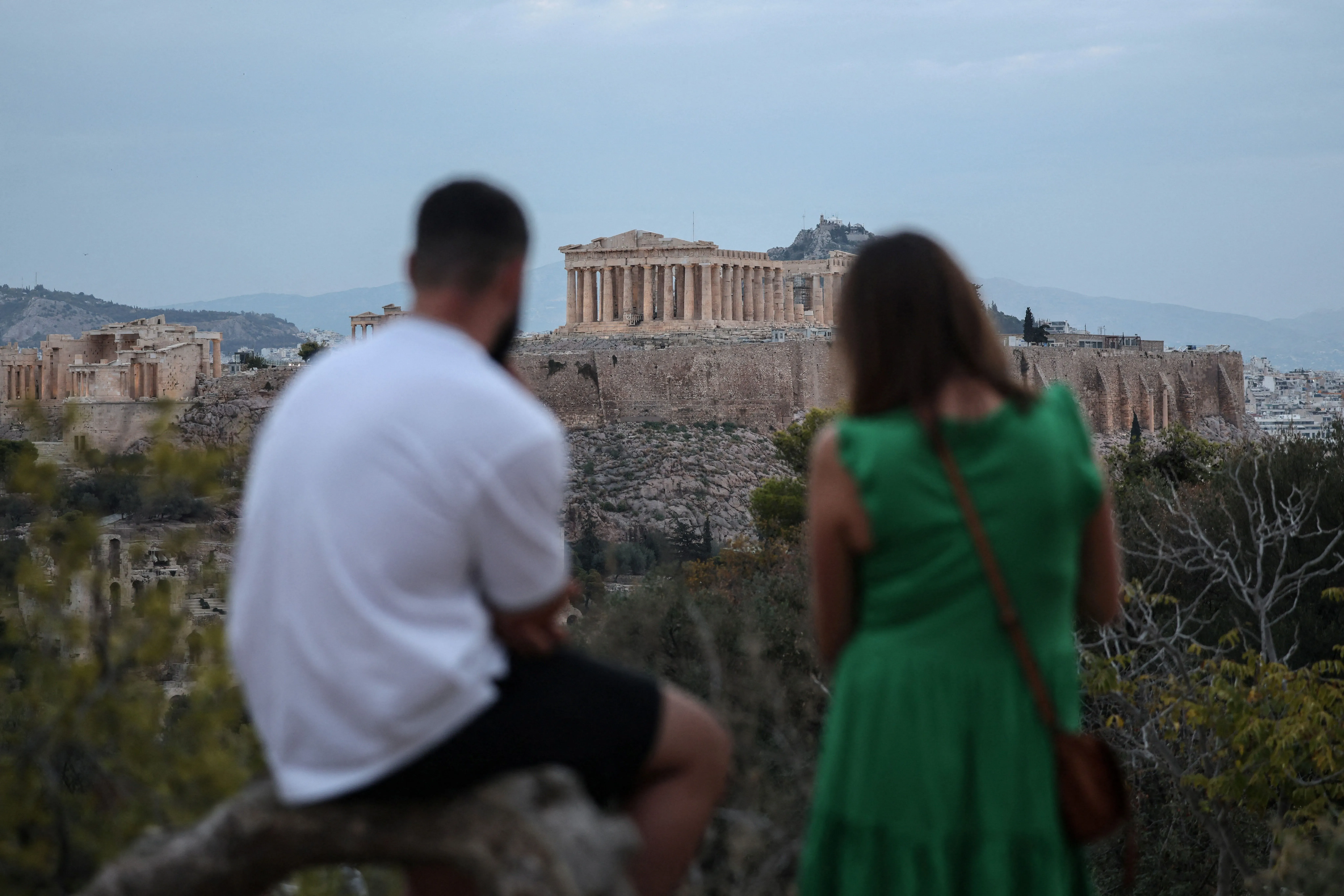 زوجان يجلسان في مقدمة الصورة، يستمتعان بإطلالة على الأكروبوليس في أثينا، مع سماء ملبدة بالغيوم في الخلفية.