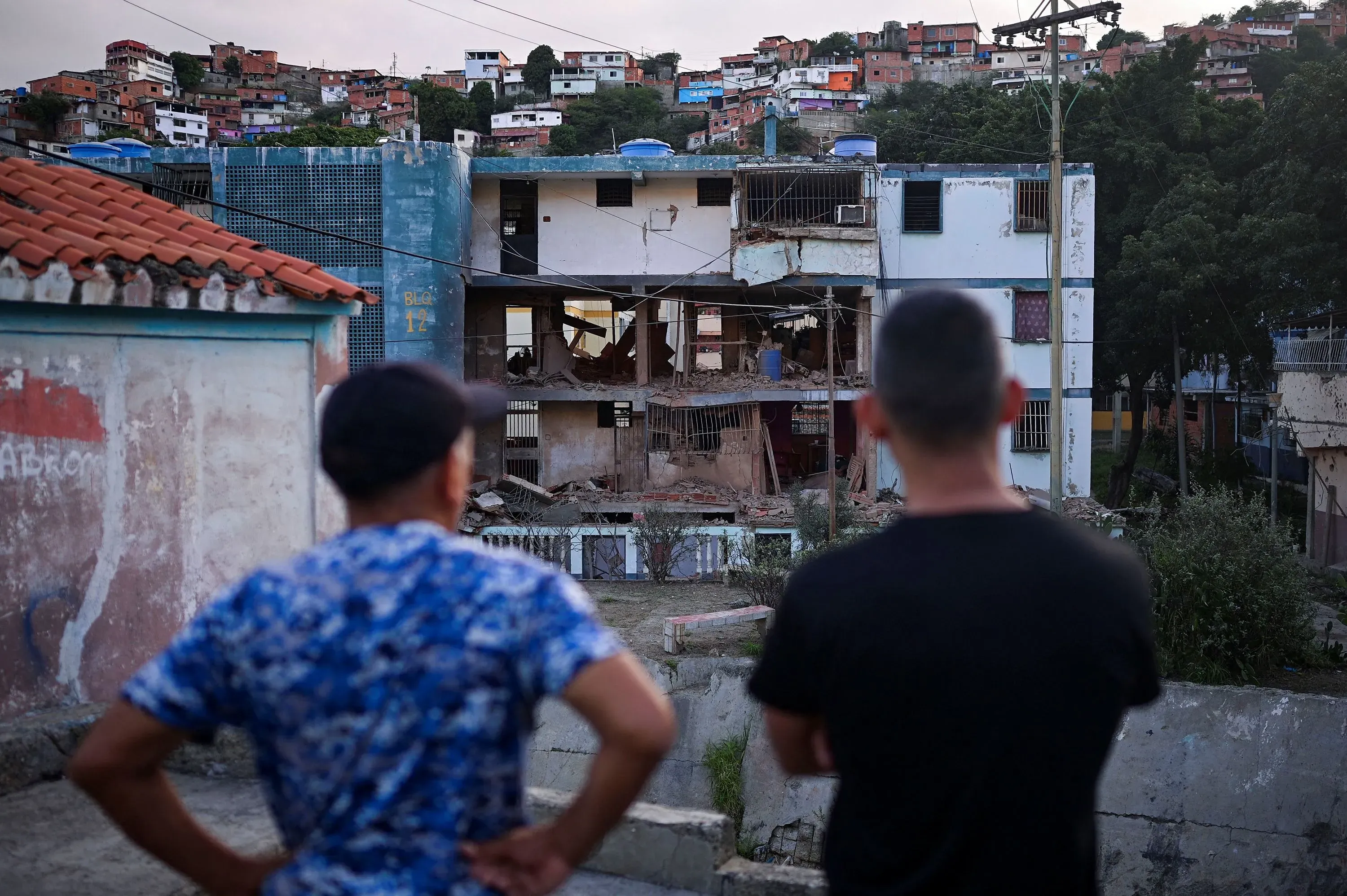 رجلان يقفان في حي شعبي في فنزويلا، ينظران إلى مبنى متضرر، مما يعكس حالة عدم اليقين بعد القبض على مادورو.