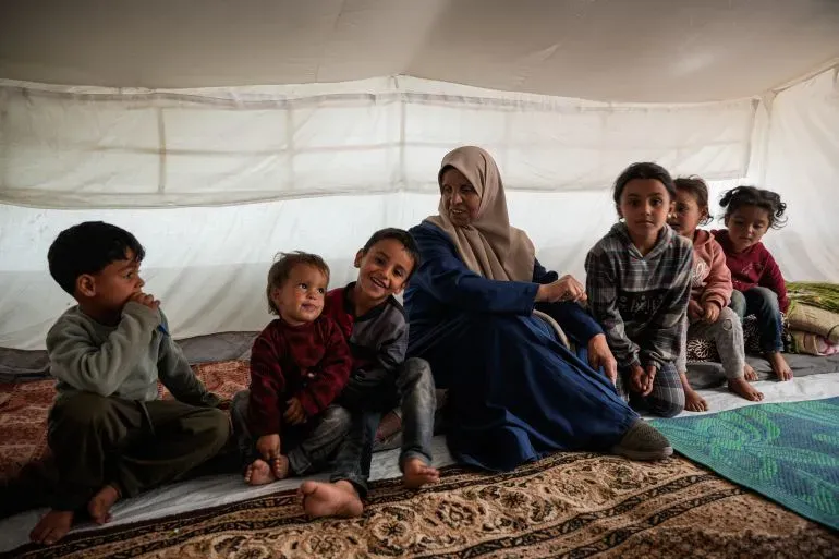 إنعام الدحدوح، امرأة مسنّة، تجلس في خيمة مع ستة من أحفادها، ممسكة بصورة لأبنائها المعتقلين، تعكس معاناتهم في ظل الحرب.