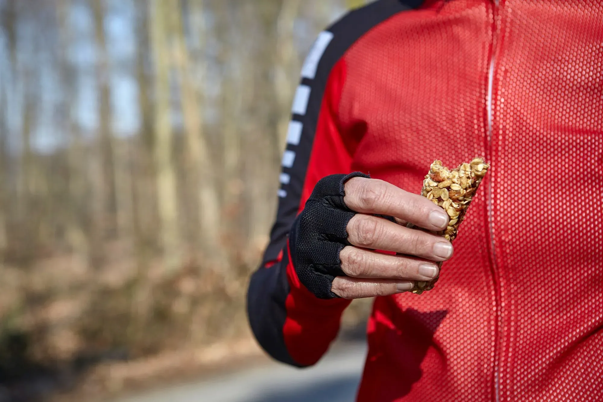 رياضي يرتدي ملابس رياضية حمراء، يحمل بار الطاقة في يده، في بيئة طبيعية، مما يعكس أهمية التغذية في الأداء الرياضي.