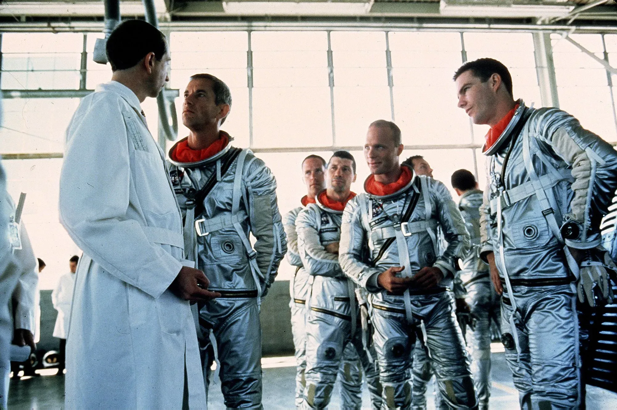 مجموعة من رواد الفضاء يرتدون بدلات فضائية فضية، يتحدثون مع شخص يرتدي معطفًا أبيض في بيئة داخلية، تعكس روح التعاون في الفضاء.