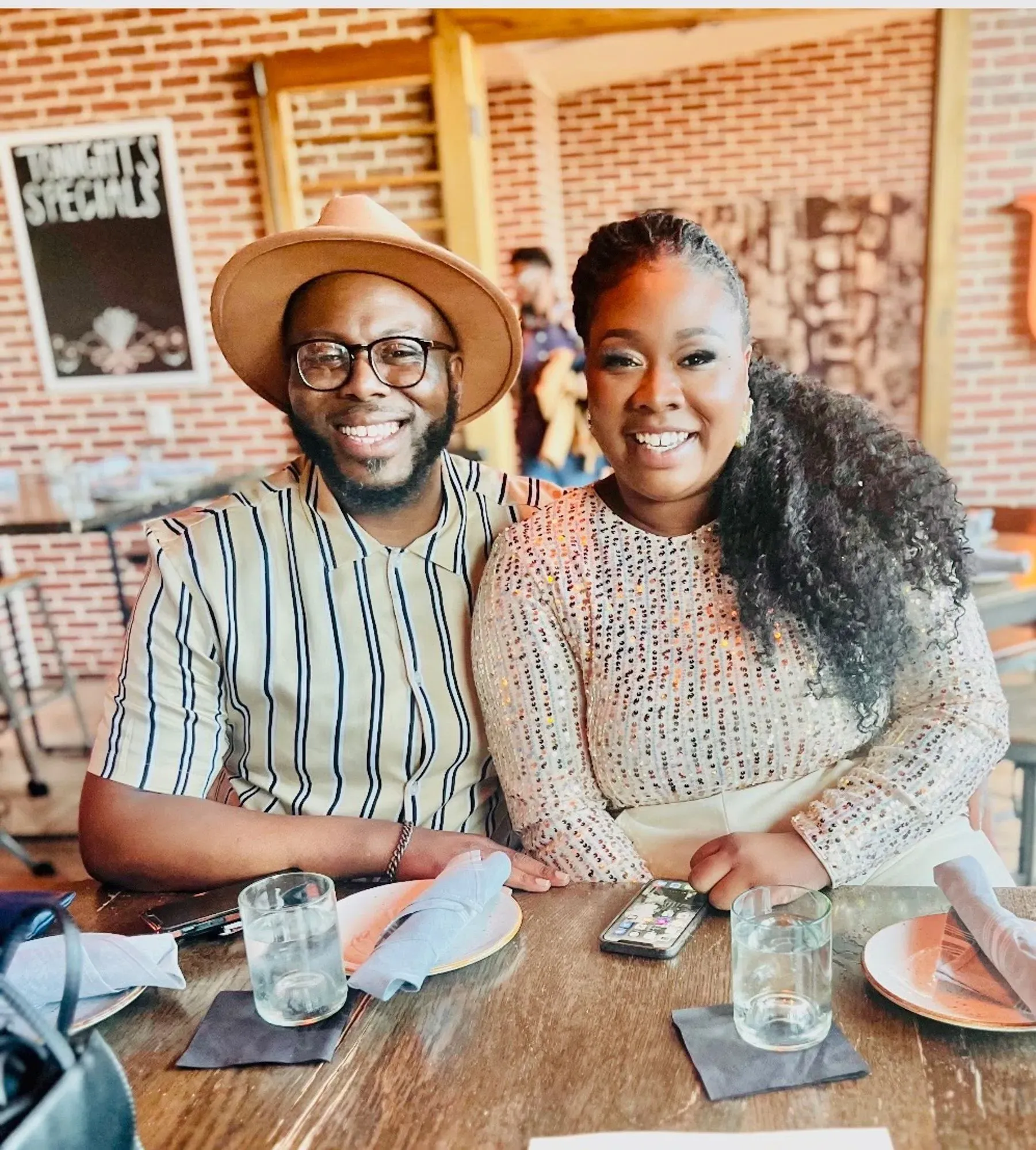 زوجان يجلسان معًا في مطعم، مبتسمين. يرتدي الرجل قميصًا مخططًا وقبعة، بينما ترتدي المرأة بلوزة لامعة.