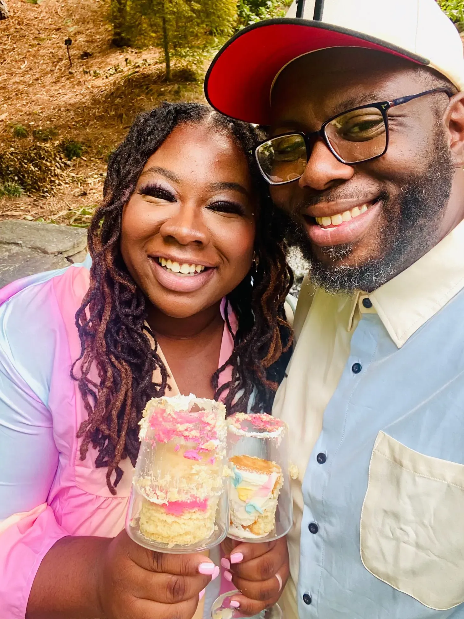 زوجان مبتسمان يحملان كعكًا في أكواب، يحتفلان بمناسبة خاصة في الهواء الطلق، مع خلفية طبيعية.