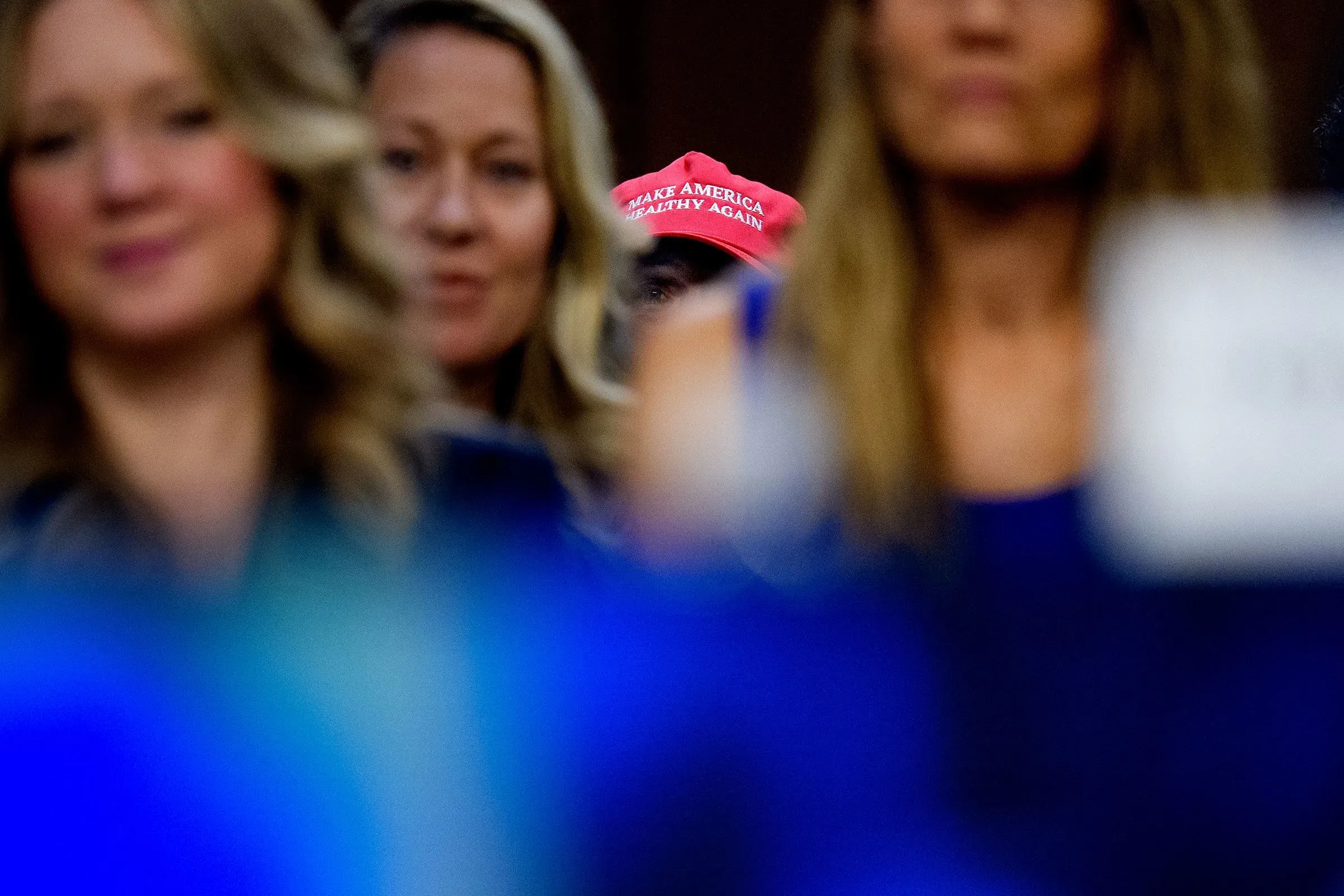 قبعة حمراء مكتوب عليها "اجعل أمريكا صحيةً من جديد" تظهر في خلفية مجموعة من النساء خلال جلسة استماع، مما يعكس الانقسامات السياسية حول سياسات الصحة العامة.