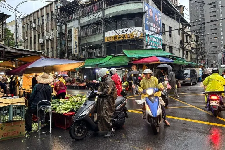 سوق مزدحم في تايوان خلال يوم ممطر، حيث يتسوق الناس ويستخدمون الدراجات النارية، مما يعكس الحياة اليومية رغم التوترات العسكرية.
