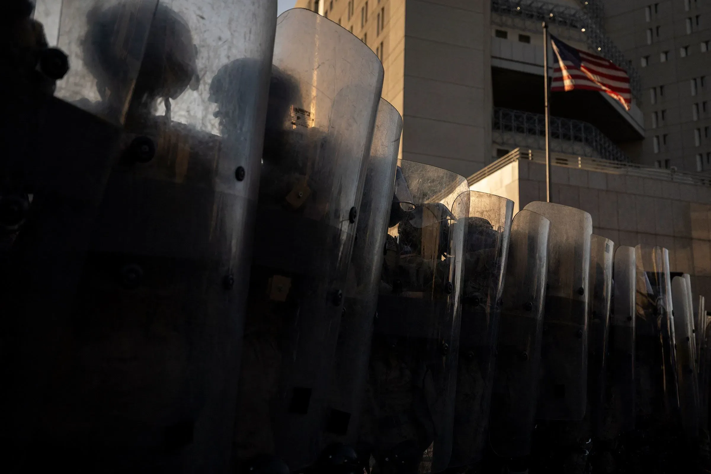 صف من الجنود يرتدون خوذات ويحملون دروعًا واقية، يقفون في صفوف أمام مبنى حكومي، مع العلم الأمريكي يرفرف في الخلفية.