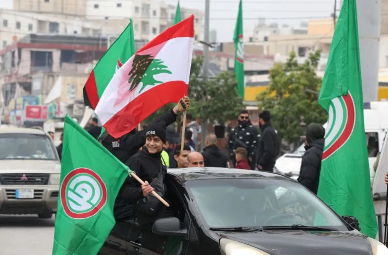 احتفال شعبي في لبنان، حيث يحمل المشاركون الأعلام اللبنانية وأعلام حزب الله، مع أجواء حماسية تعكس الانقسامات السياسية في البلاد.