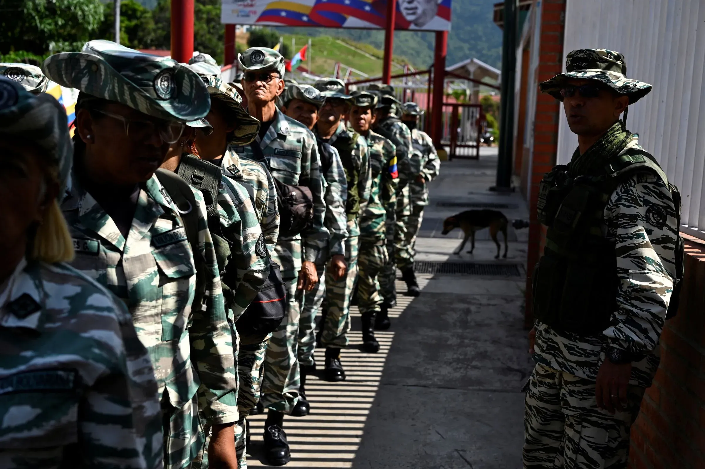 جنود فنزويليين يرتدون زيًا عسكريًا متنوعًا، يقفون في صف طويل في موقع عسكري، مع خلفية تظهر أعلام وطنية وصورة لقائد سياسي.