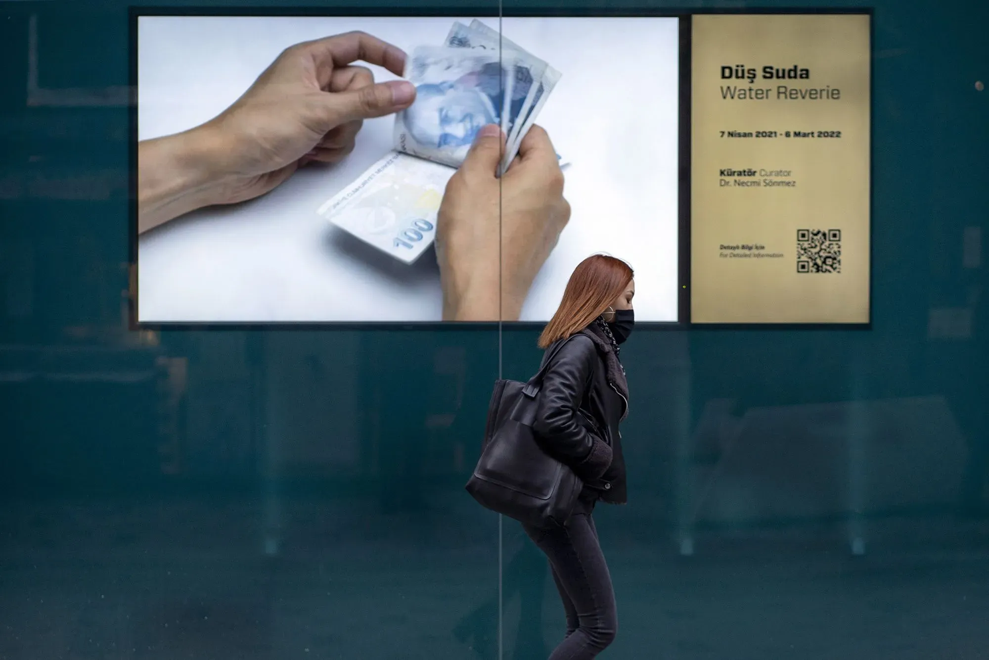 امرأة ترتدي سترة سوداء تسير بجانب لوحة إعلانات تعرض يدين تحملان نقودًا تركية، تعكس قضايا التضخم والسياسة النقدية في تركيا.