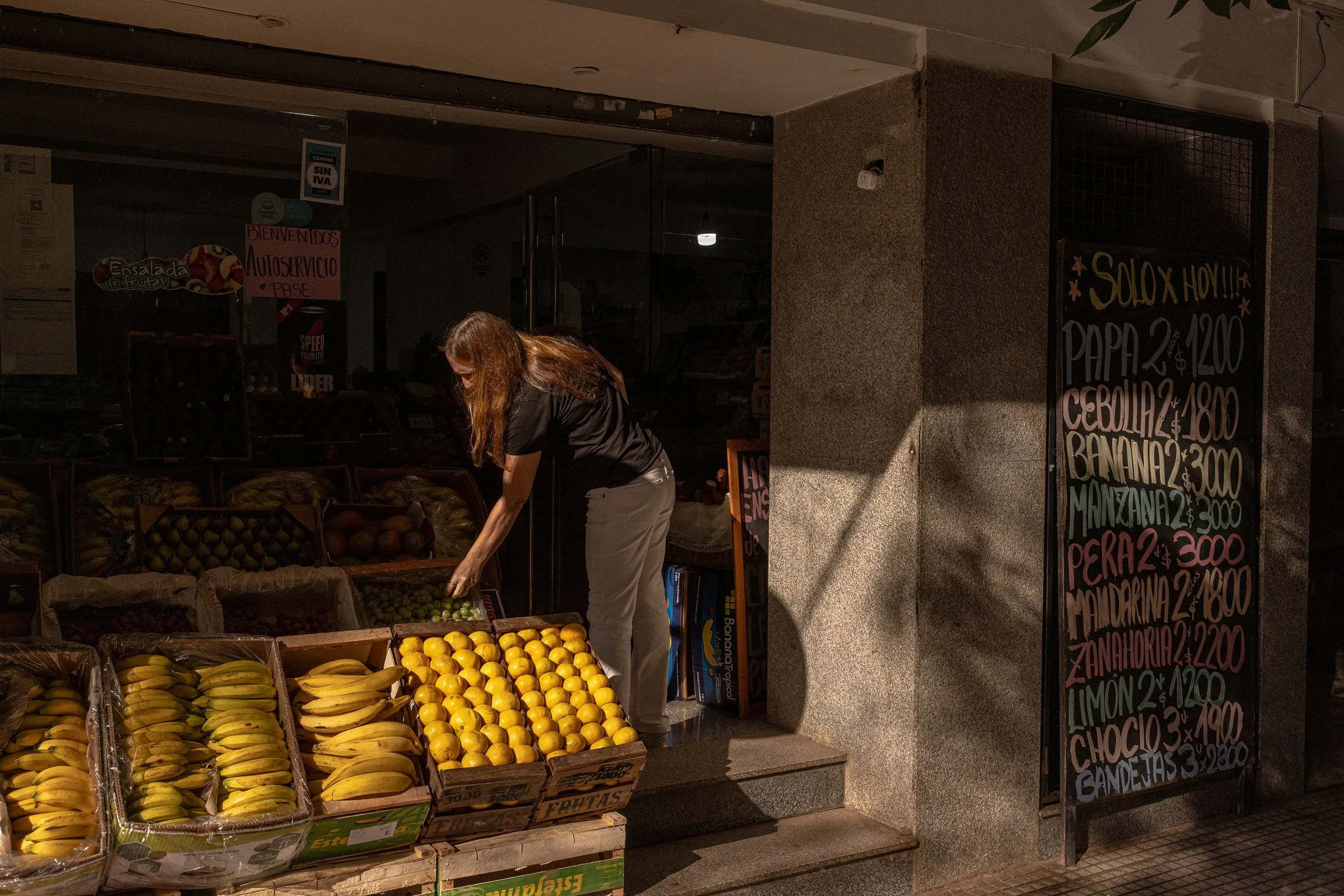 امرأة ترتب الفواكه في متجر محلي، مع قائمة أسعار مكتوبة على لوح بجانبها، تعكس الوضع الاقتصادي الحالي.