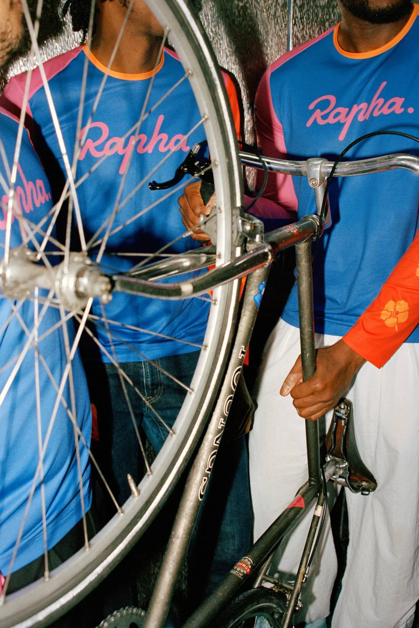 شبان يرتدون قمصان Rapha الزرقاء والوردية يحملون دراجة هوائية سوداء في مشهد يعكس التعاونات الفنية لعام 2024.