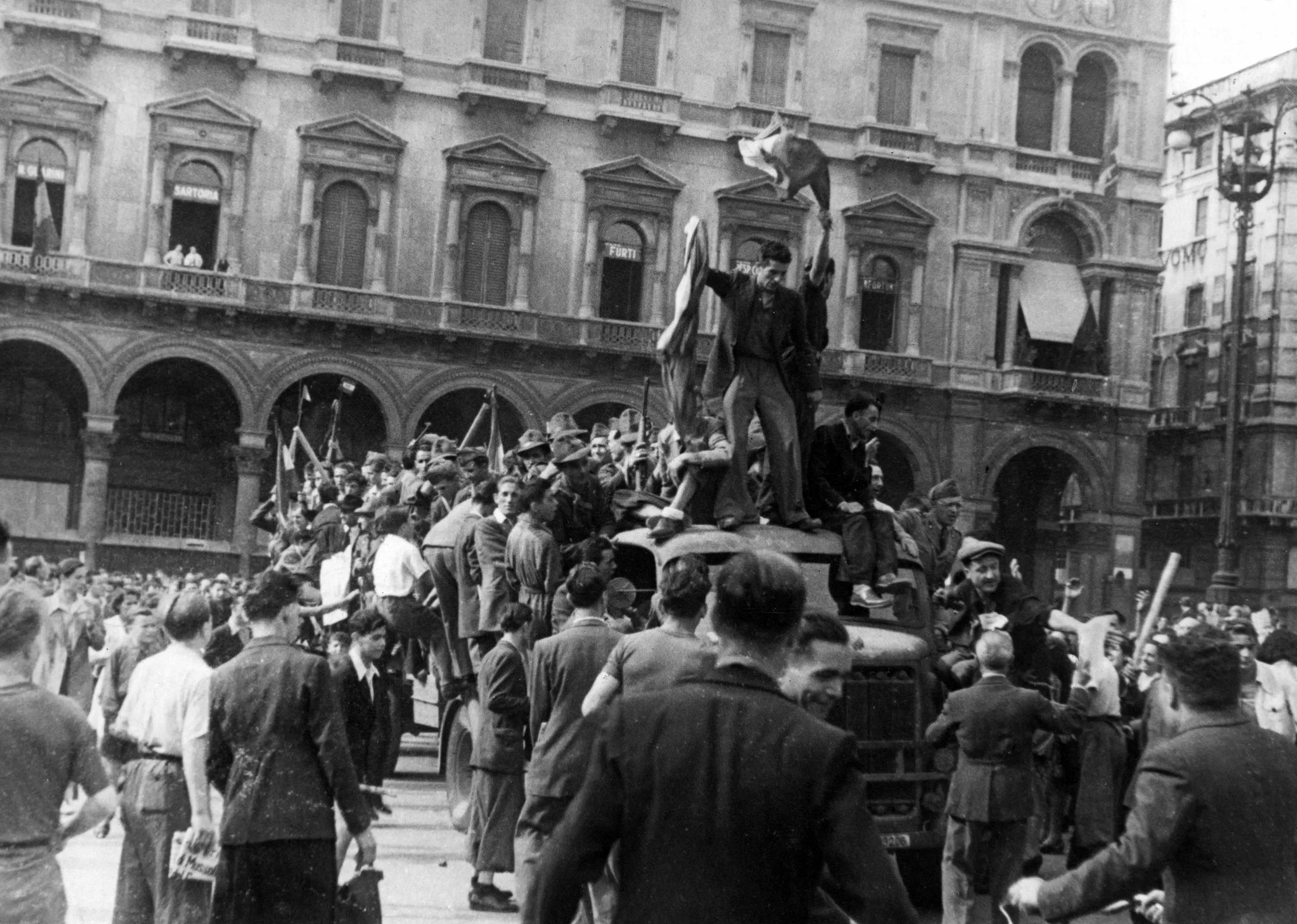 مظاهرة تاريخية في ساحة عامة، حيث يتجمع حشد كبير من الناس حول سيارة عسكرية، مع رفع الأعلام والاحتجاجات.