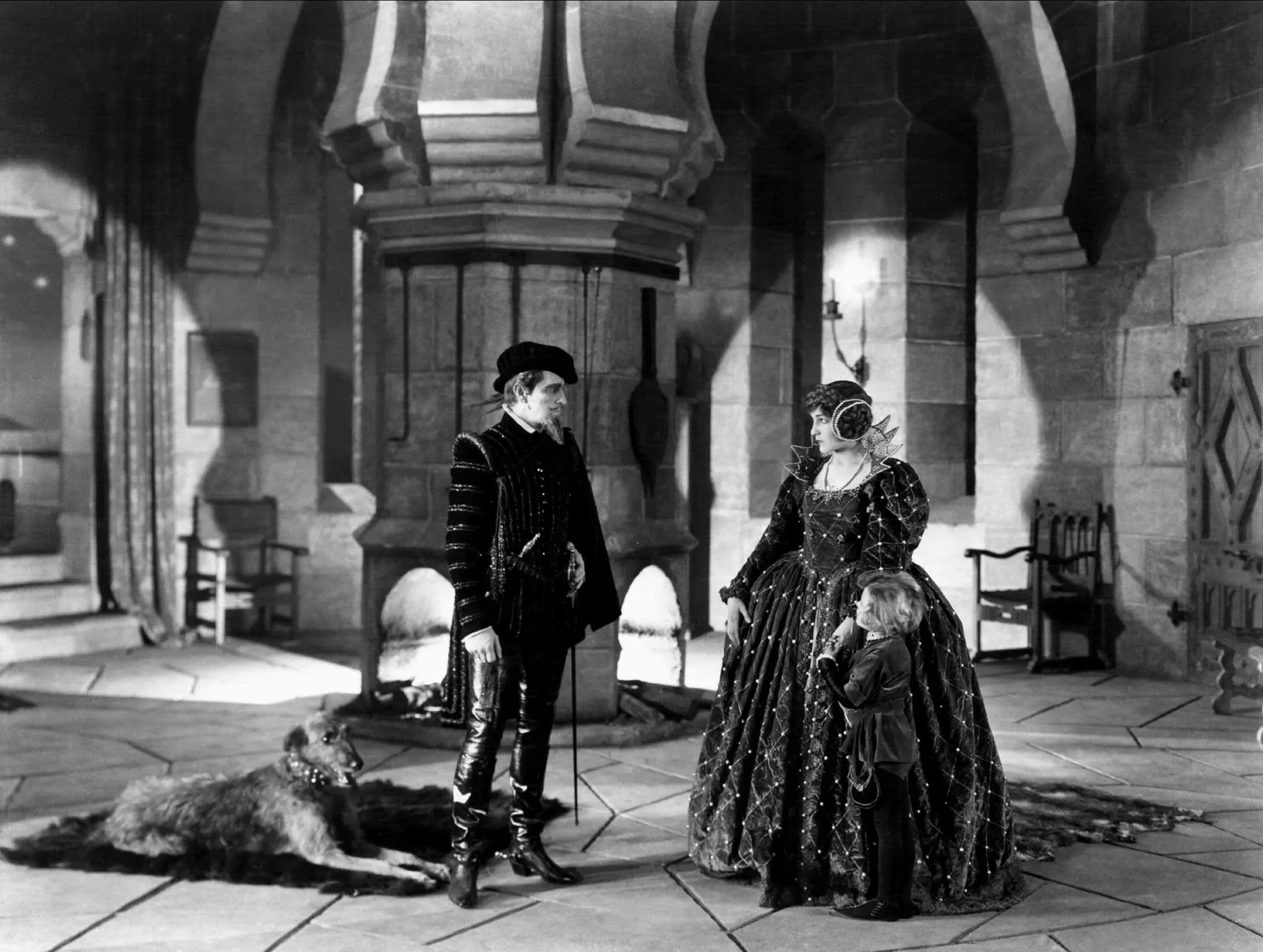 مشهد من فيلم درامي قديم يظهر شخصيات ترتدي أزياء تاريخية في غرفة كبيرة، مع وجود كلب على الأرض، مما يعكس تاريخ السينما الكلاسيكية.