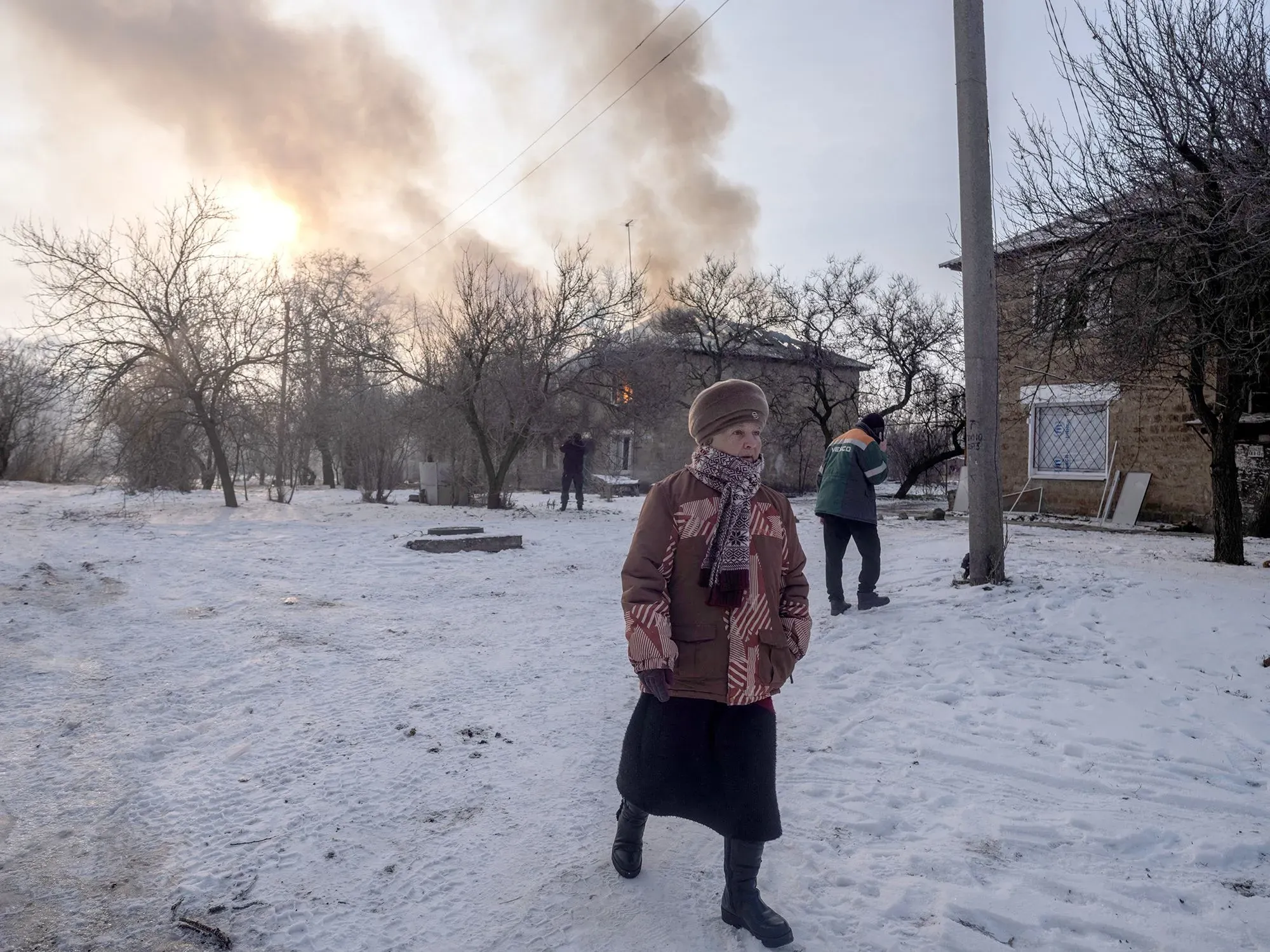 امرأة ترتدي ملابس دافئة تسير في منطقة مغطاة بالثلوج، بينما تظهر أعمدة الدخان تتصاعد من منزل محترق في الخلفية، مما يعكس آثار النزاع في دونباس.