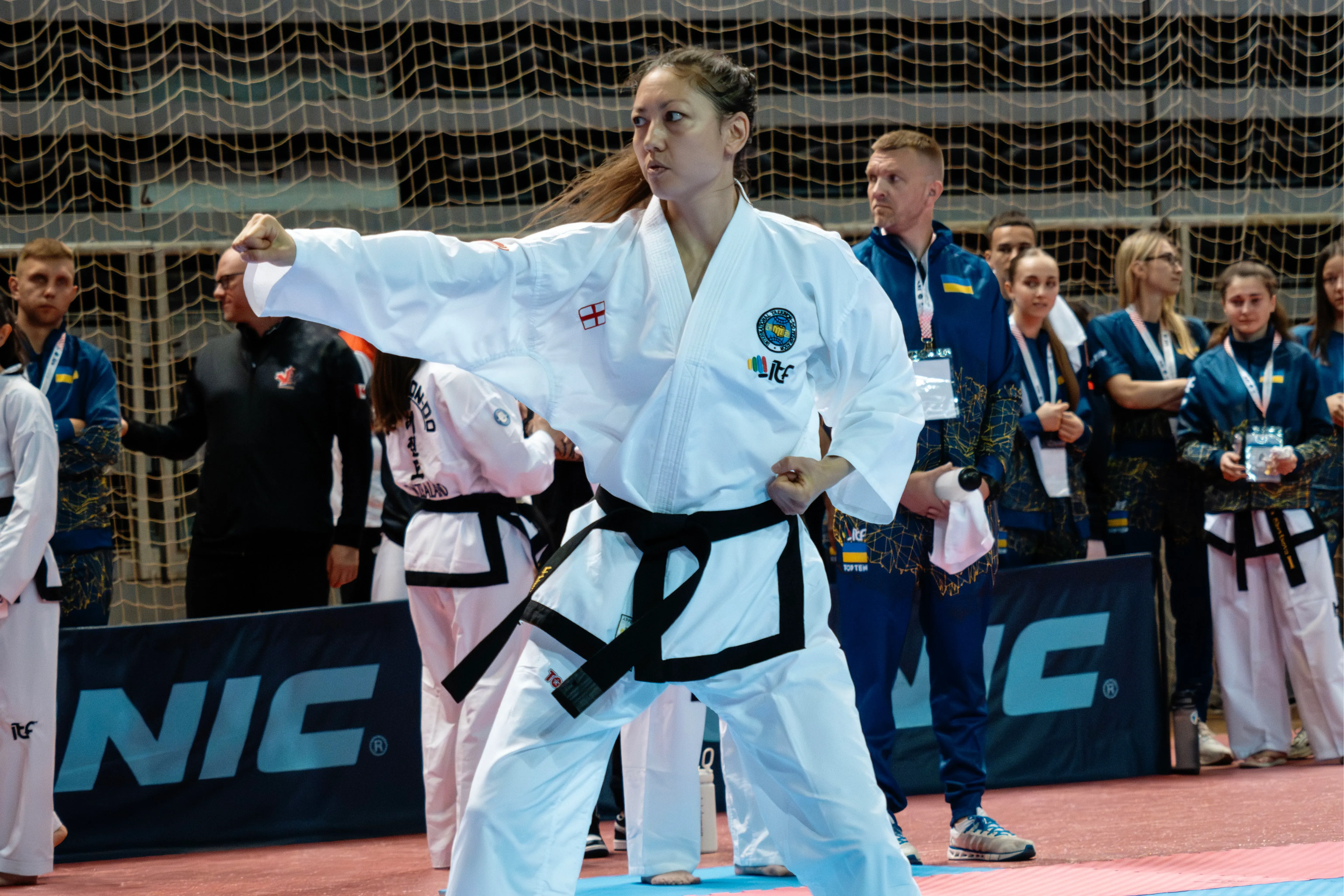 لاعبة تايكوندو ترتدي زيًا أبيض مع حزام أسود، تؤدي حركة قتالية في بطولة، بينما يتابعها فريقها من الخلف.