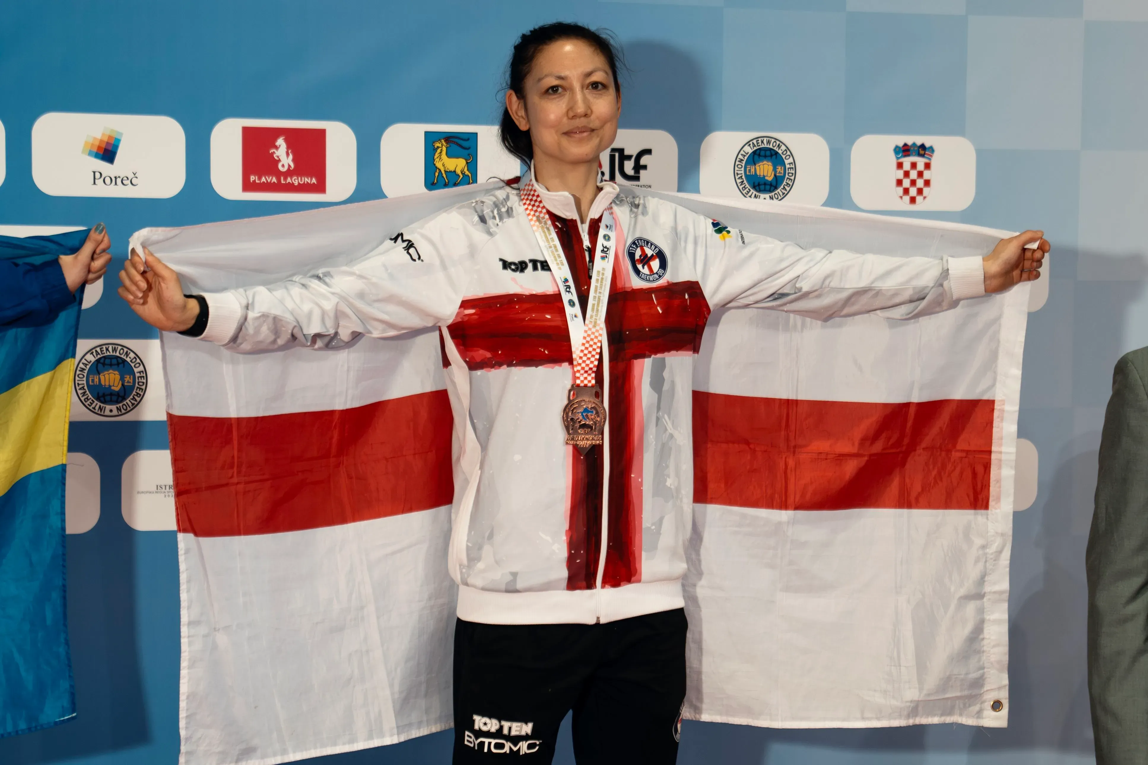 رياضية تحمل علم إنجلترا بفخر بعد الفوز بميدالية في بطولة التايكوندو، تعكس إنجازات الفريق الوطني الإنجليزي.