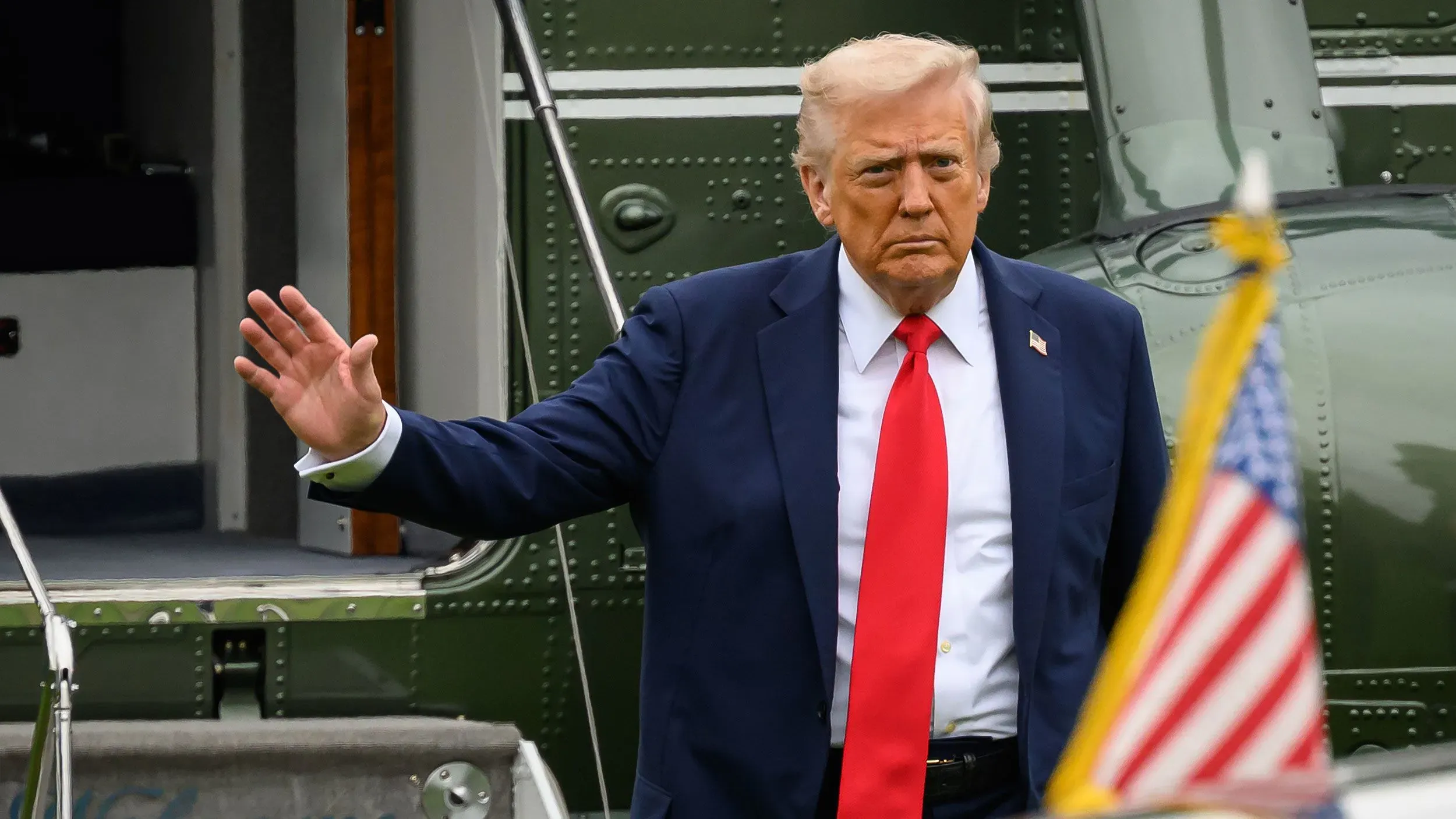 الرئيس السابق دونالد ترامب يسير نحو المروحية، مرتديًا بدلة زرقاء وقميص أبيض ربطة عنق حمراء، مع العلم الأمريكي في المقدمة.