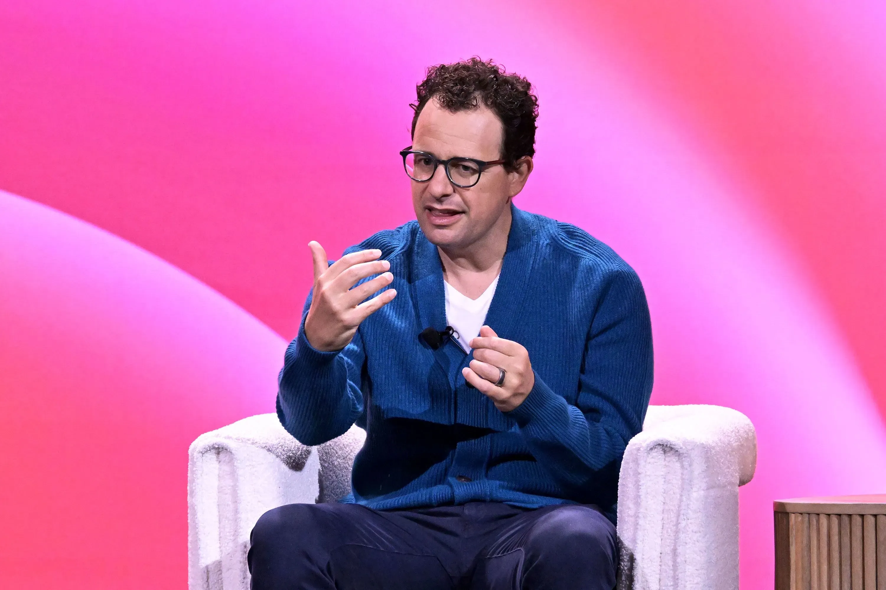 رجل يتحدث في حدث تقني، يرتدي سترة زرقاء، مع خلفية ملونة، يعكس قضايا السلامة والنمو في مجال الذكاء الاصطناعي.