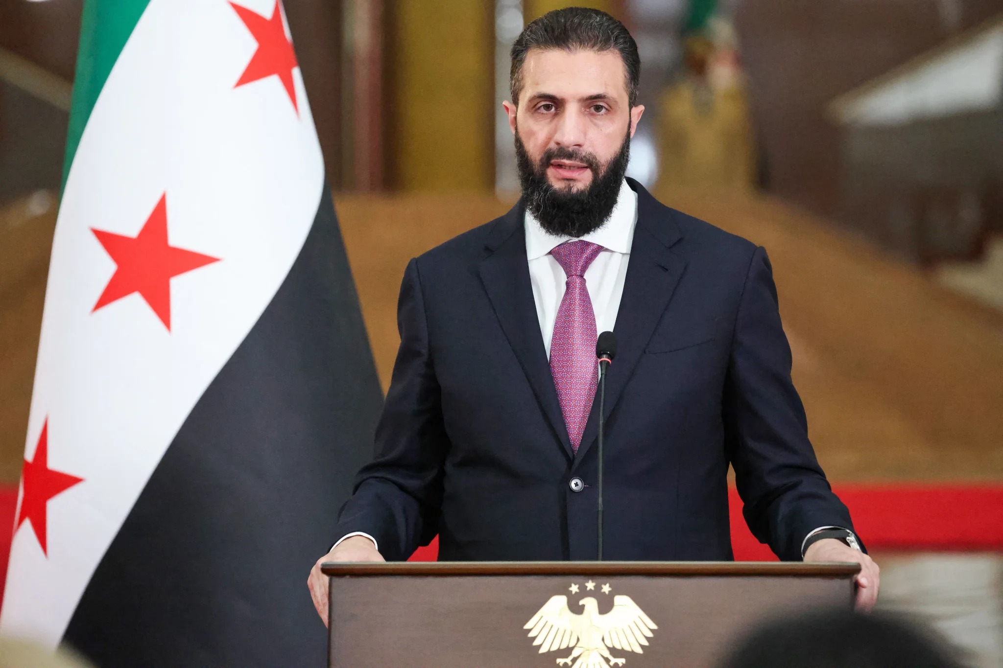 رئيس سوريا أحمد الشرع يخاطب الجمهور من خلف منبر رسمي وعلم الثورة السورية خلفه، في سياق تعزيز العدالة الانتقالية بعد سقوط نظام الأسد.
