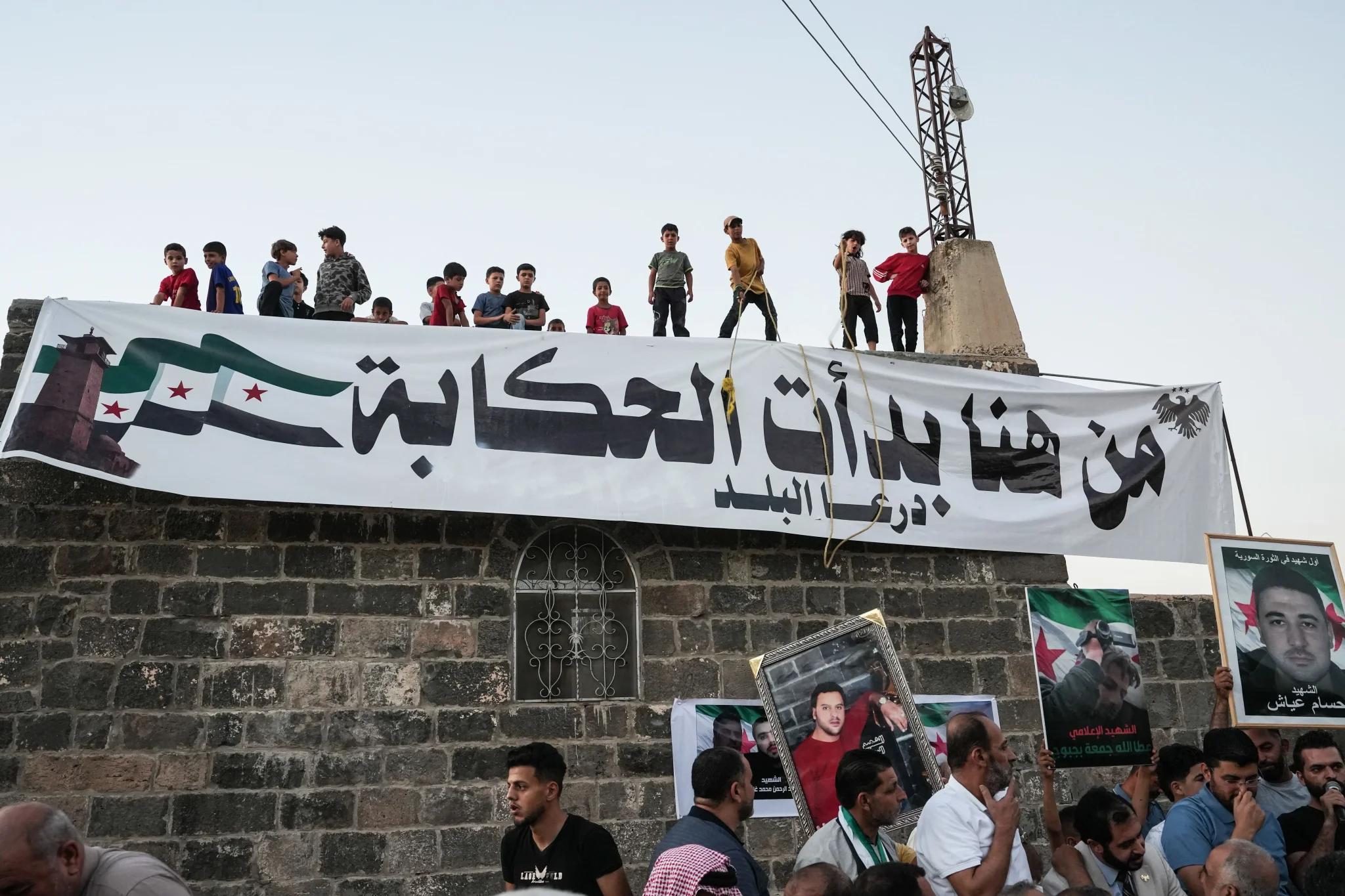 أطفال وسكان يحتفلون في درعا برفع لافتة كتب عليها "من هنا بدأت الحكاية" تعبيراً عن بداية مرحلة العدالة الانتقالية في سوريا بعد حكم الإعدام على الأسد.