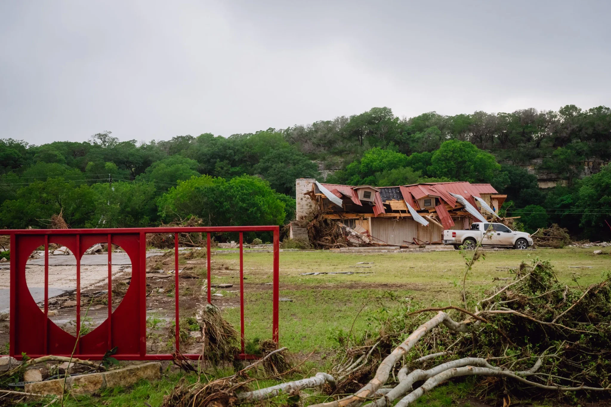 منزل متضرر وجدار حديدي أحمر عليه شكل قلب وسط آثار فيضان نهر Guadalupe في Texas Hill Country مع وجود شاحنة بيضاء.