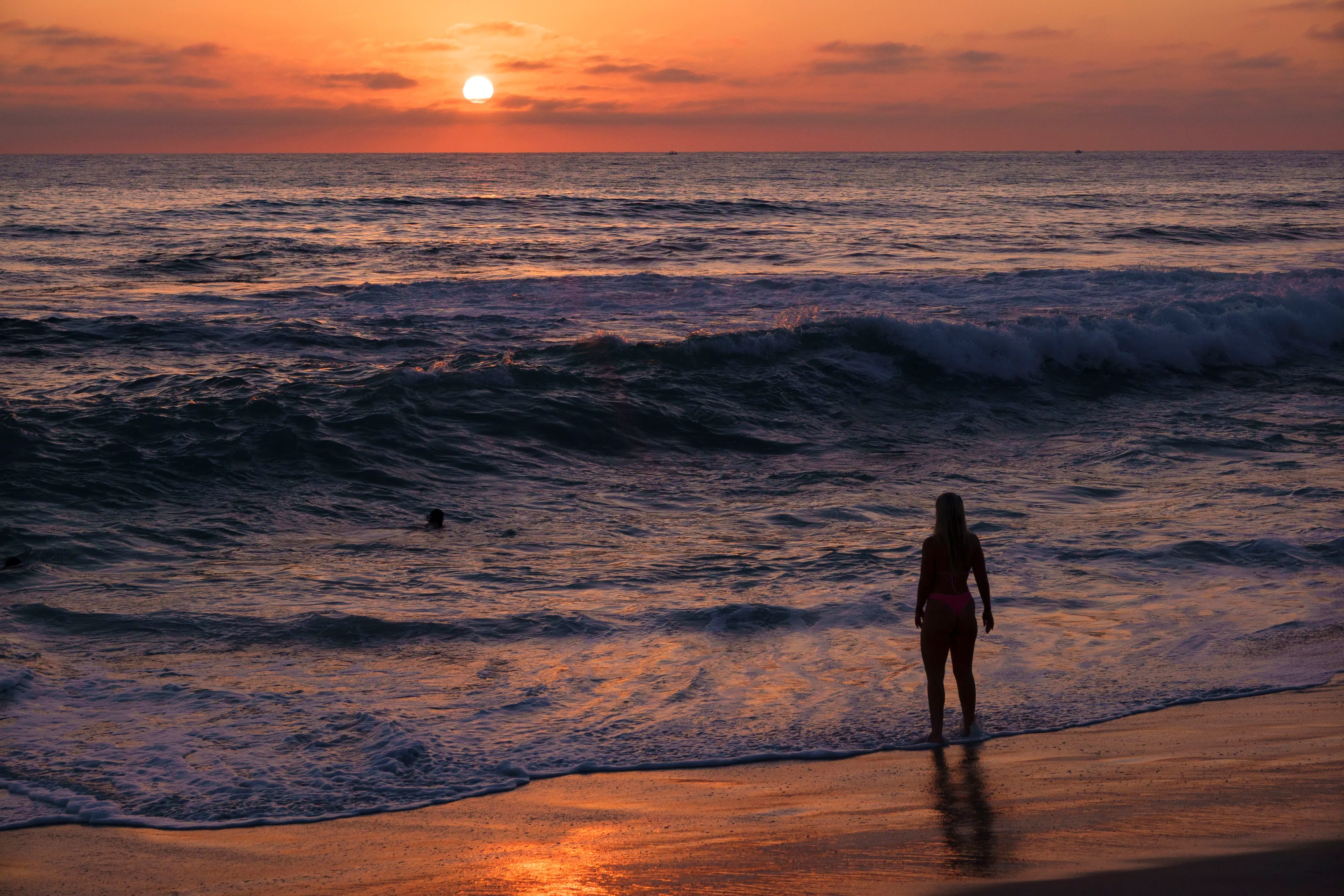 غروب الشمس يضيء سماء ساحل المحيط الهادئ، مع سيدة واقفة على الشاطئ أمام الأمواج، مما يعكس تأثير الحرارة البحرية على الطقس.