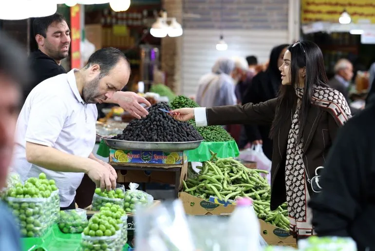 امرأة تشتري التمر من بائع في سوق طهران وسط ارتفاع التضخم وتأثير الحصار الأمريكي على الاقتصاد الإيراني.