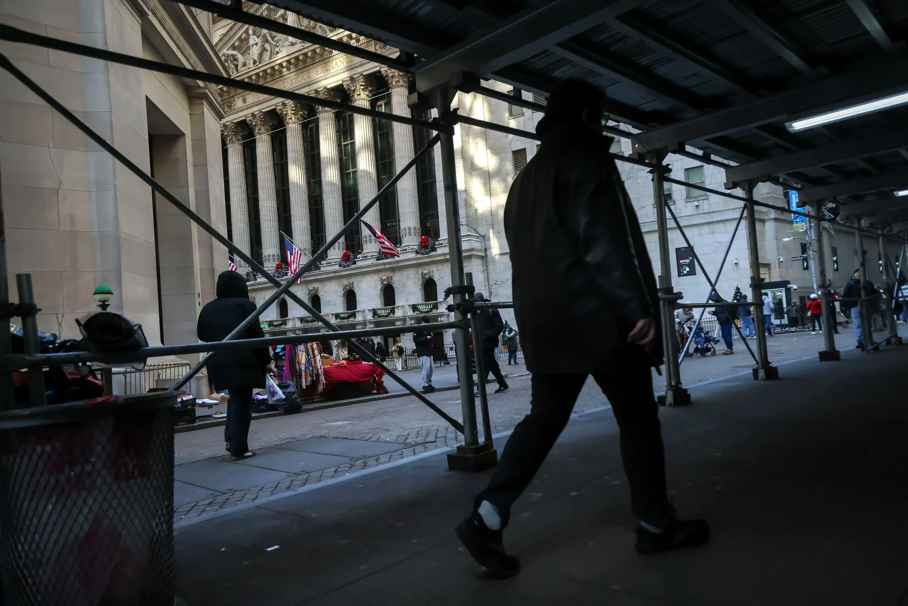 شخص يسير تحت سقالات بالقرب من بورصة نيويورك، مع وجود أعلام أمريكية معلقة، مما يعكس النشاط الاقتصادي في المنطقة.