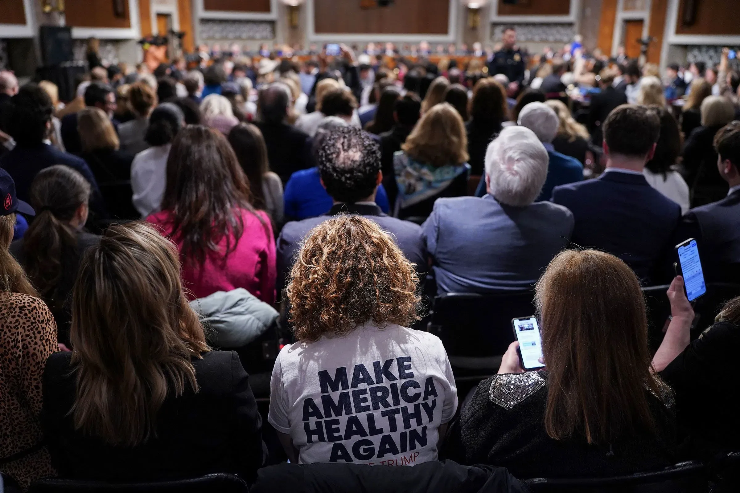 امرأة تجلس في تجمع عام، ترتدي قميصًا مكتوبًا عليه "اجعل أمريكا صحية مرة أخرى"، بينما تستمع إلى مناقشات حول لقاحات الأطفال.