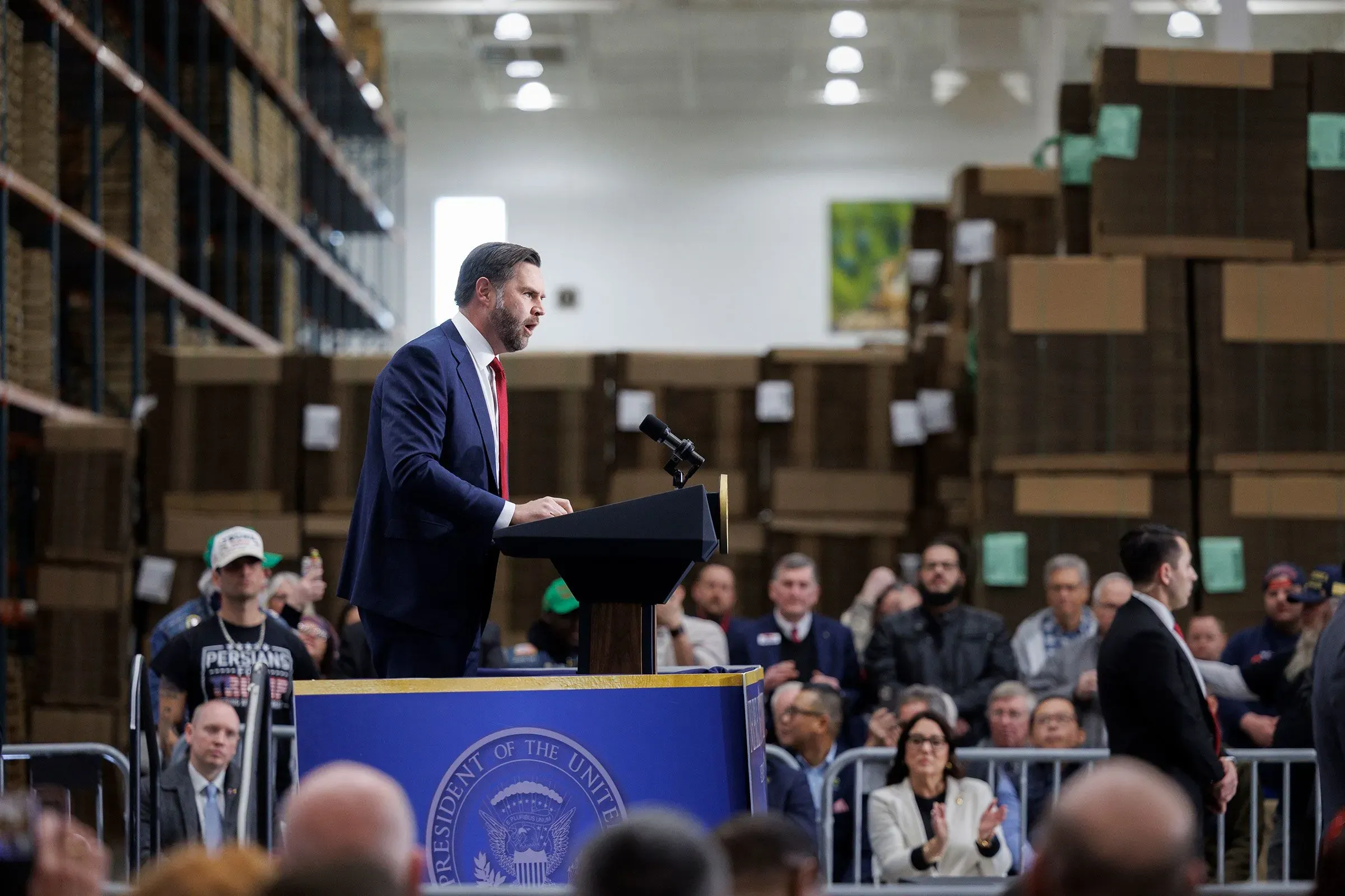 نائب الرئيس جي دي فانس يلقي خطابًا في تجمع جماهيري، مع خلفية من صناديق التخزين، حيث يستعرض قضايا الاقتصاد وتأثيرها على الأمريكيين.