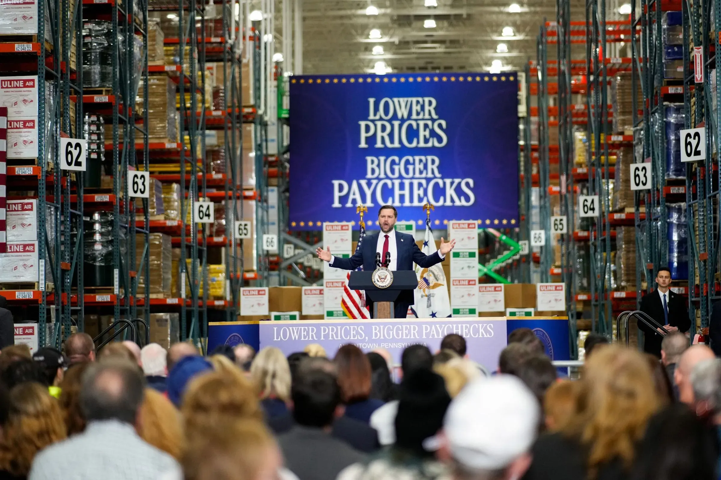 نائب الرئيس فانس يتحدث أمام حشد في مستودع، مع لافتة تروج لأسعار أقل ورواتب أكبر، مما يعكس قضايا الاقتصاد الحالية.