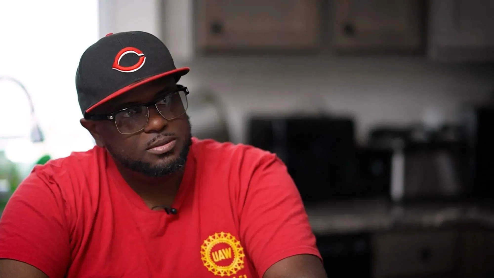 رجل يرتدي قميصًا أحمر وقبعة سوداء، يجلس في مطبخ، يعبر عن آرائه حول الوضع السياسي والاقتصادي في الولايات المتحدة.