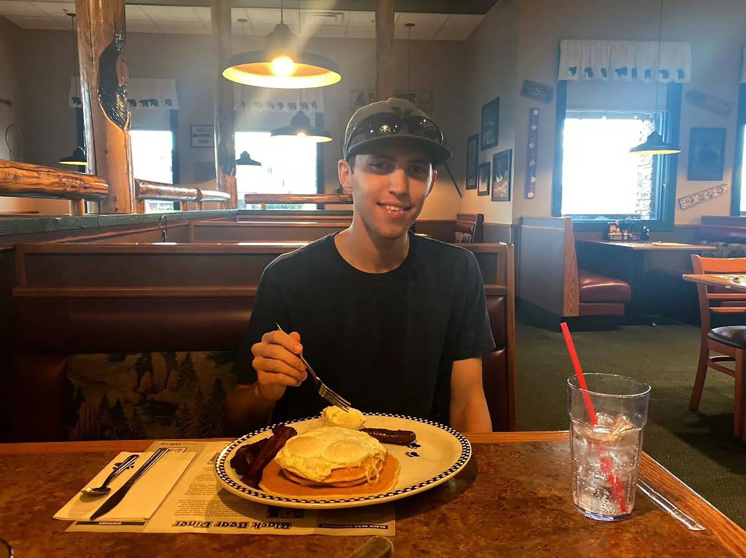 شاب يجلس في مطعم مع طبق من الفطور يتضمن البيض ولحم الخنزير المقدد، مبتسمًا أمام الكاميرا.