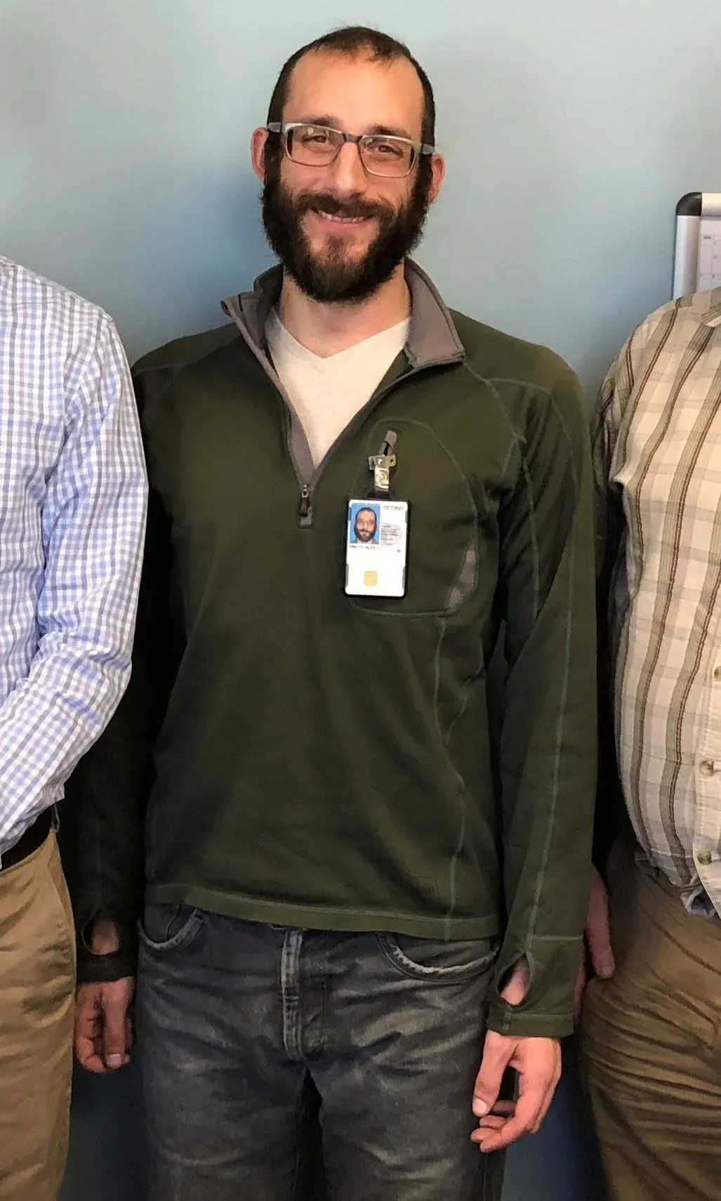 رجل يرتدي نظارات وقميصًا أخضر، مبتسمًا، يقف بين شخصين آخرين. تُظهر الصورة أجواء غير رسمية، مع خلفية بسيطة.