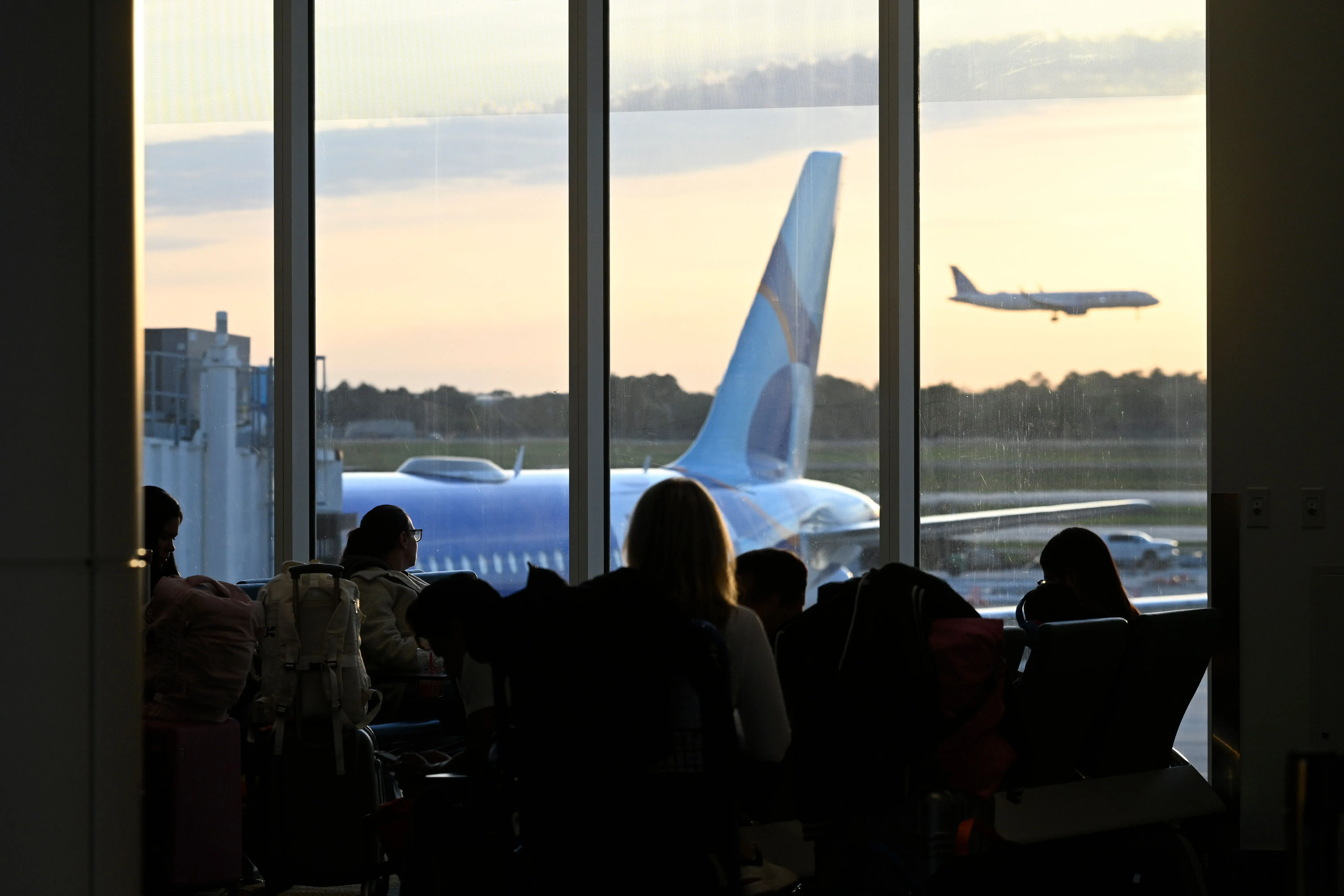 مشهد لمطار مزدحم حيث يجلس المسافرون في منطقة الانتظار، مع طائرة تقلع في الخلفية، مما يعكس الازدحام وتأثير نقص موظفي إدارة أمن النقل.