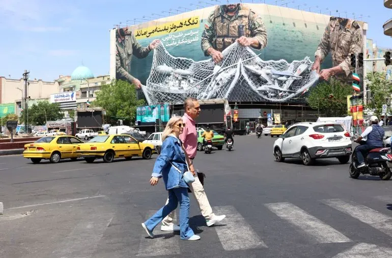 امرأة ورجل يعبران ممرًا للمشاة في مدينة إيرانية، مع وجود لوحة إعلانات كبيرة خلفهما تُظهر مشهدًا لصيد الأسماك.